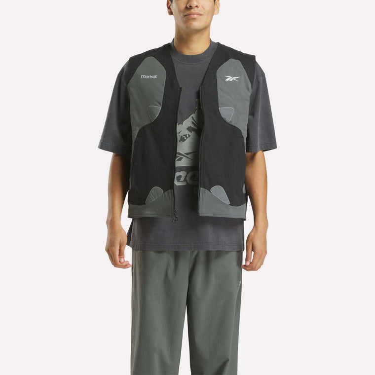 A person stands facing forward against a white background, wearing a loose dark gray Reebok T-shirt with a graphic logo, gray pants, and the Reebok x MARKET Reversible Vest in black and gray—capturing modern streetwear style with hands by their sides.
