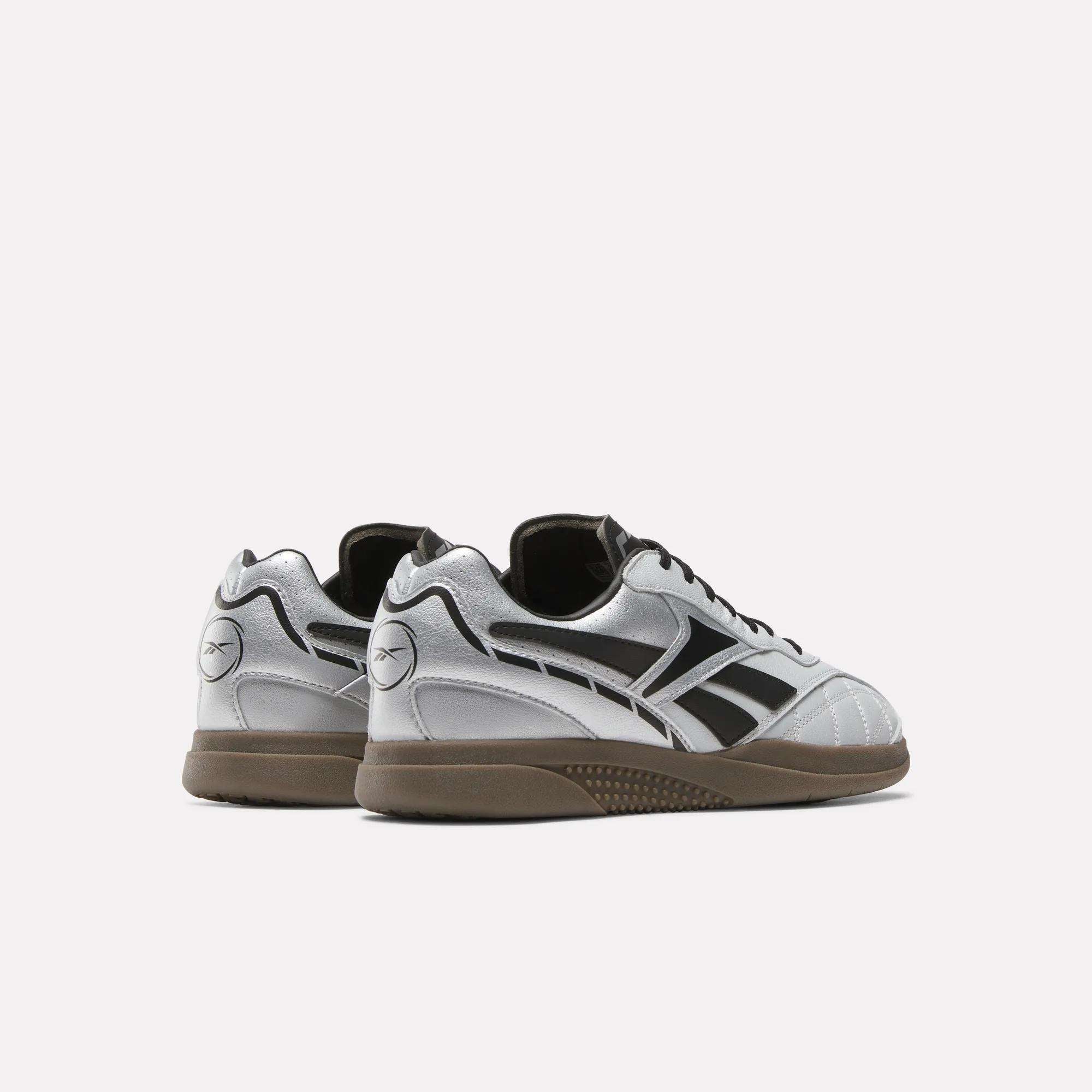 The Reebok Hammer Street Shoes in silver and black feature dark soles, black logo accents, full-grain leather panels, and detailed stitching. Pictured from the back and side against a white background, these sleek shoes offer a soccer-inspired look.