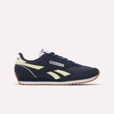 The Women's Classic AZ Shoes by Reebok feature a navy blue upper, pale yellow side stripes, white midsole, and gum outsole for a vintage look. Reebok branding is on the side, tongue, and heel. The sneaker is shown from the side on a white background.
