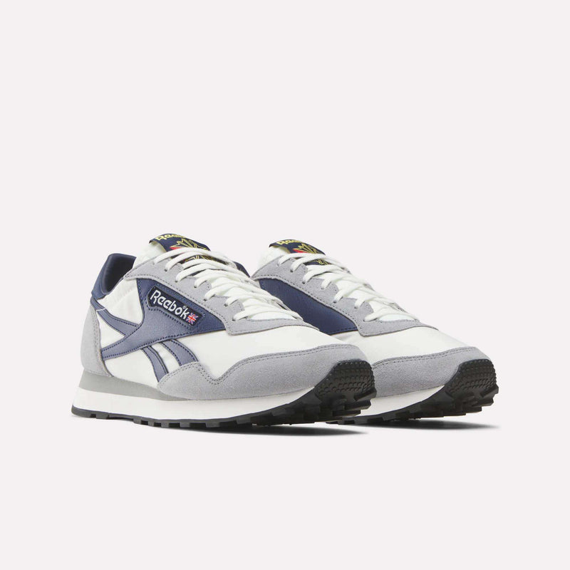 A pair of Reebok Aztec II Shoes in white, grey, and navy with black outsoles. Inspired by retro running and ’80s style, they feature the Reebok logo and side stripes on a plain white background.