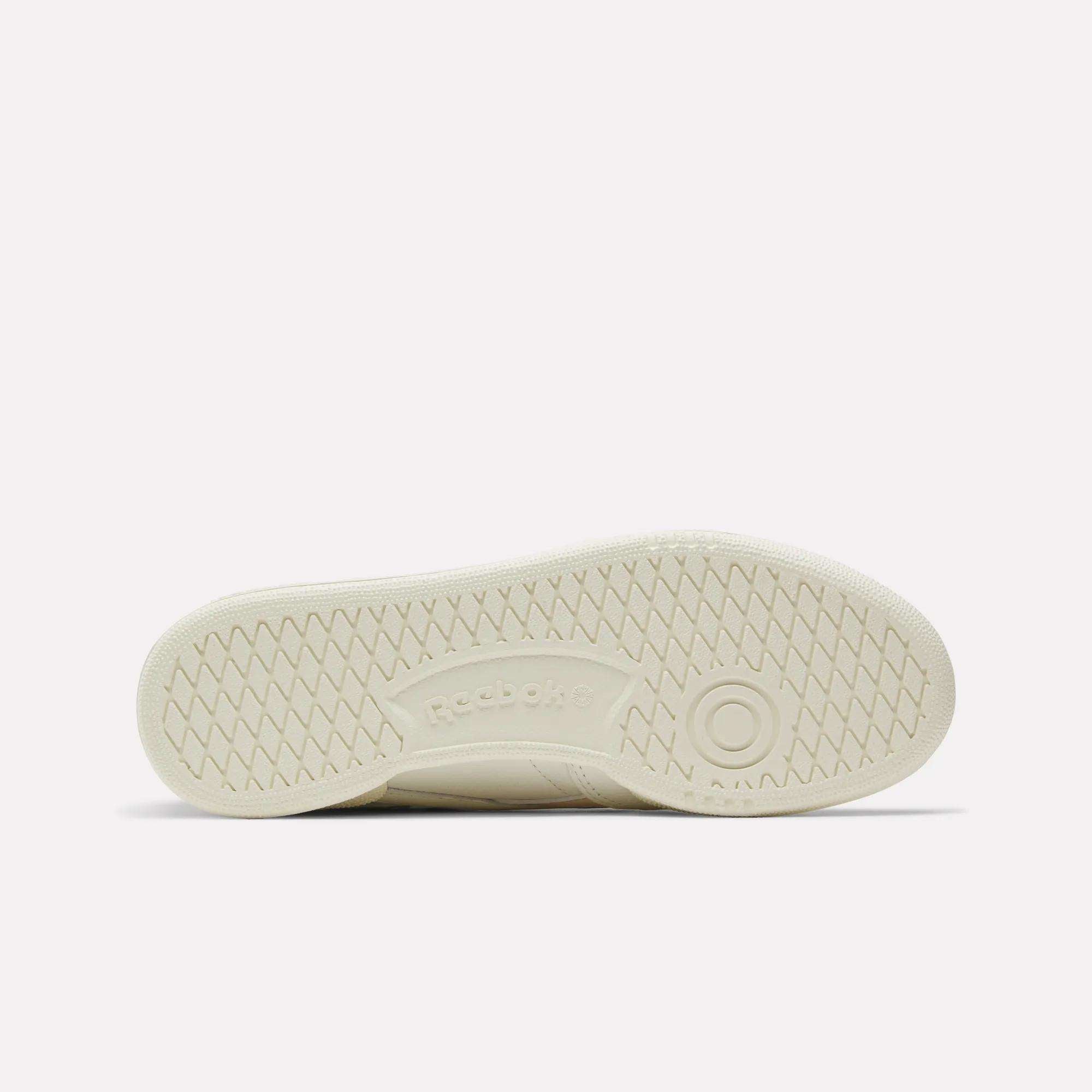 The image shows the bottom view of a beige Reebok Women's Club C Revenge Vintage Shoe sole, highlighting a herringbone tread, Reebok logo at the center, and a circular design near the heel—classic vintage-inspired details.