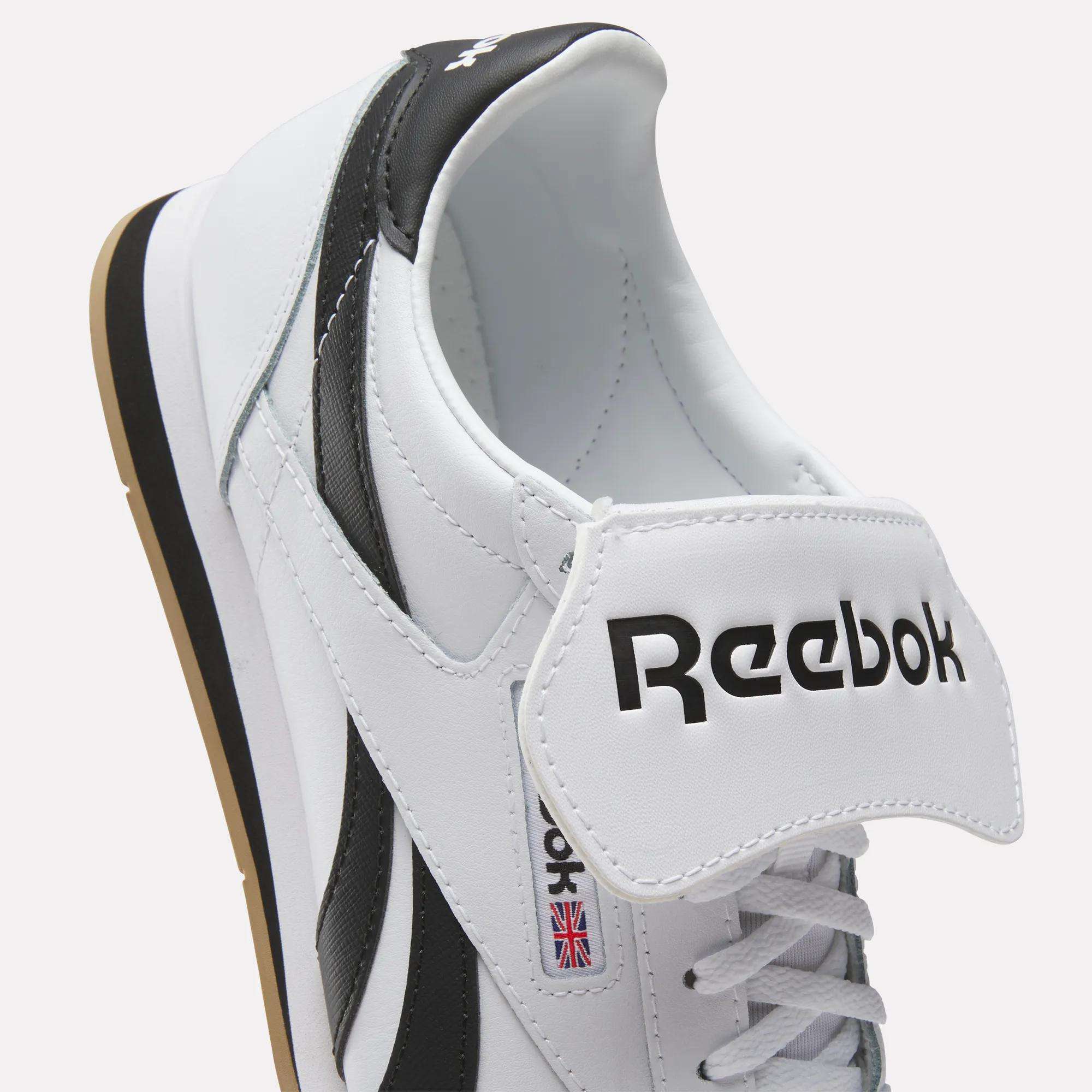 A close-up of white leather Reebok Campio XT Kilty Shoes with black accents, showcasing the Reebok logo on the tongue and a small Union Jack flag on the side. The tennis-inspired style stands out with the tongue pulled forward, revealing the inside.