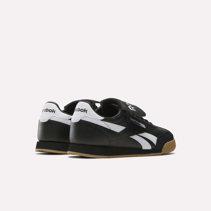 A pair of Reebok Campio XT Kilty Shoes in black with white stripes, white heel accents, gum soles, and velcro straps are displayed on a light background, angled to showcase the back and side tennis-inspired design.