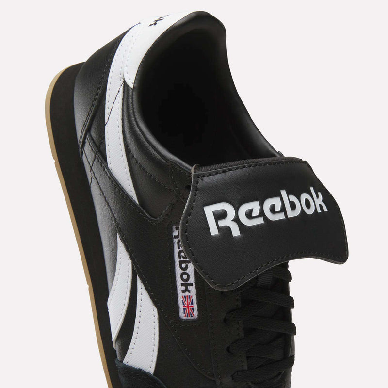 A close-up of the Reebok Campio XT Kilty Shoes in black leather with white stripes, featuring tennis-inspired style, a bold Reebok logo on a large tongue flap, and a small UK flag tag on the side.