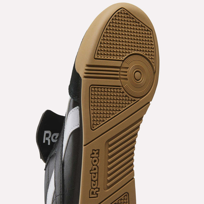 Close-up of the Reebok Campio XT Kilty Shoes' sole, featuring a textured brown rubber outsole with circular and rectangular patterns, the Reebok logo, and part of the black and white upper for a tennis-inspired look.