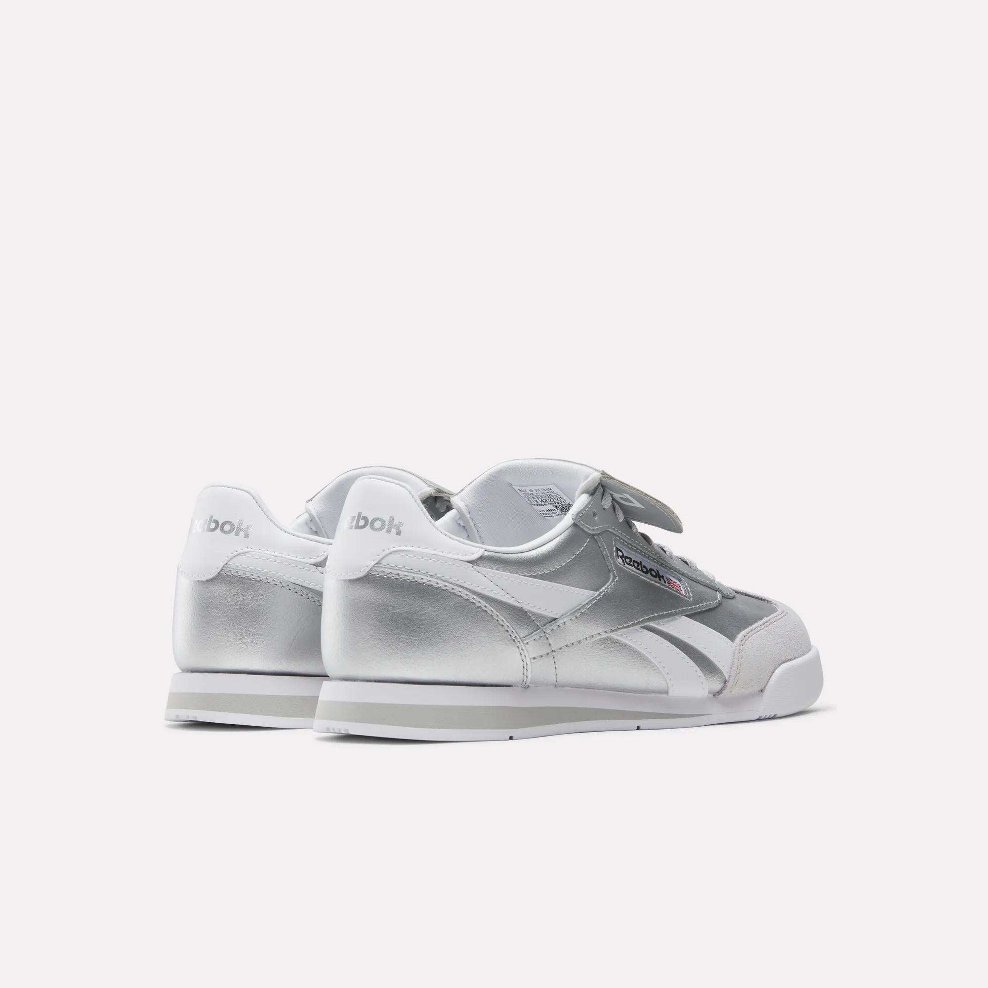 A pair of Reebok Campio XT Kilty Shoes in silver and white, shown from the back and side, with the Reebok logo visible on the heel and side. These tennis-inspired sneakers are displayed against a plain white background.