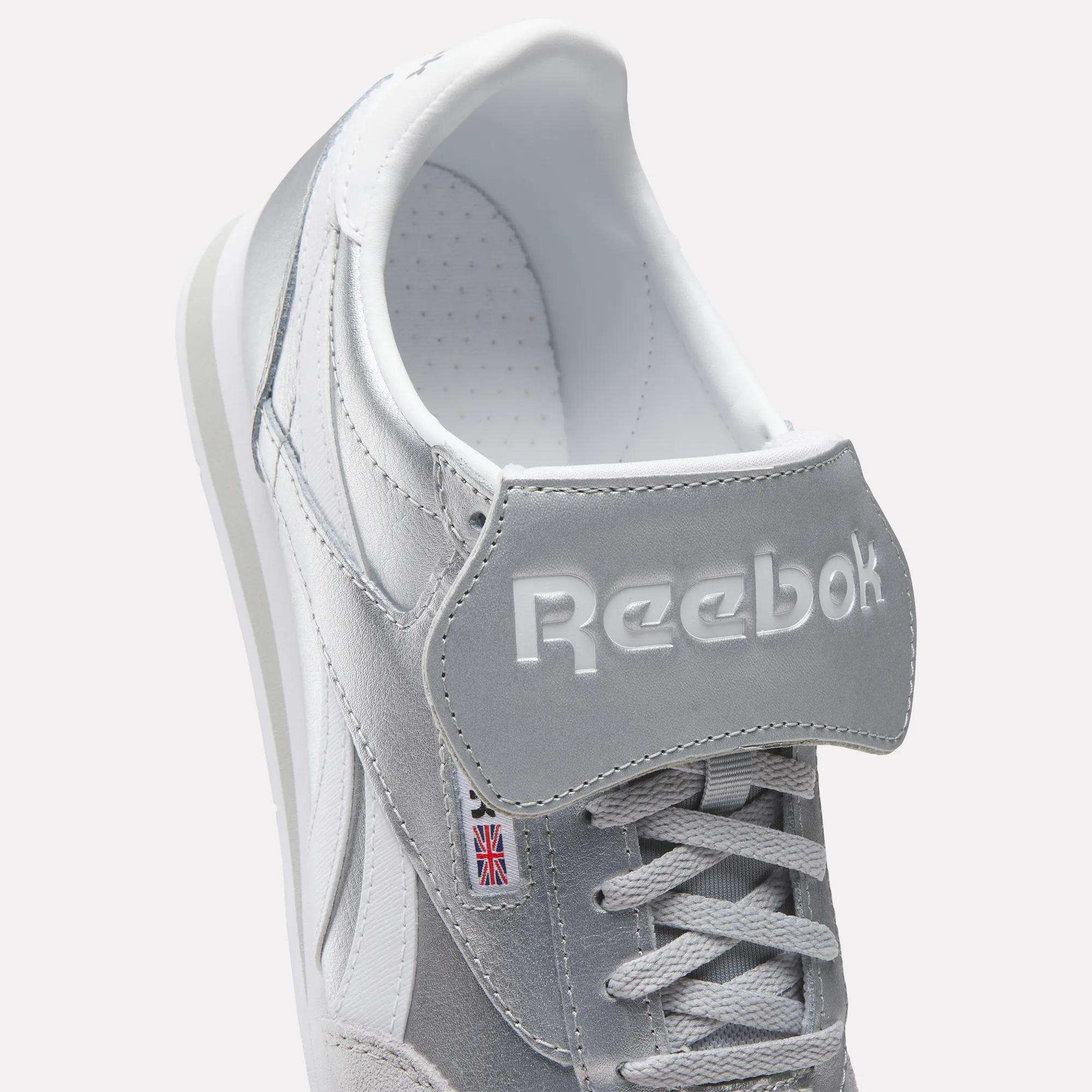 Close-up of Reebok Campio XT Kilty Shoes in gray and white with white laces, embossed Reebok logo on the tongue, and a small Union Jack label on the side, highlighting their classic tennis-inspired style.