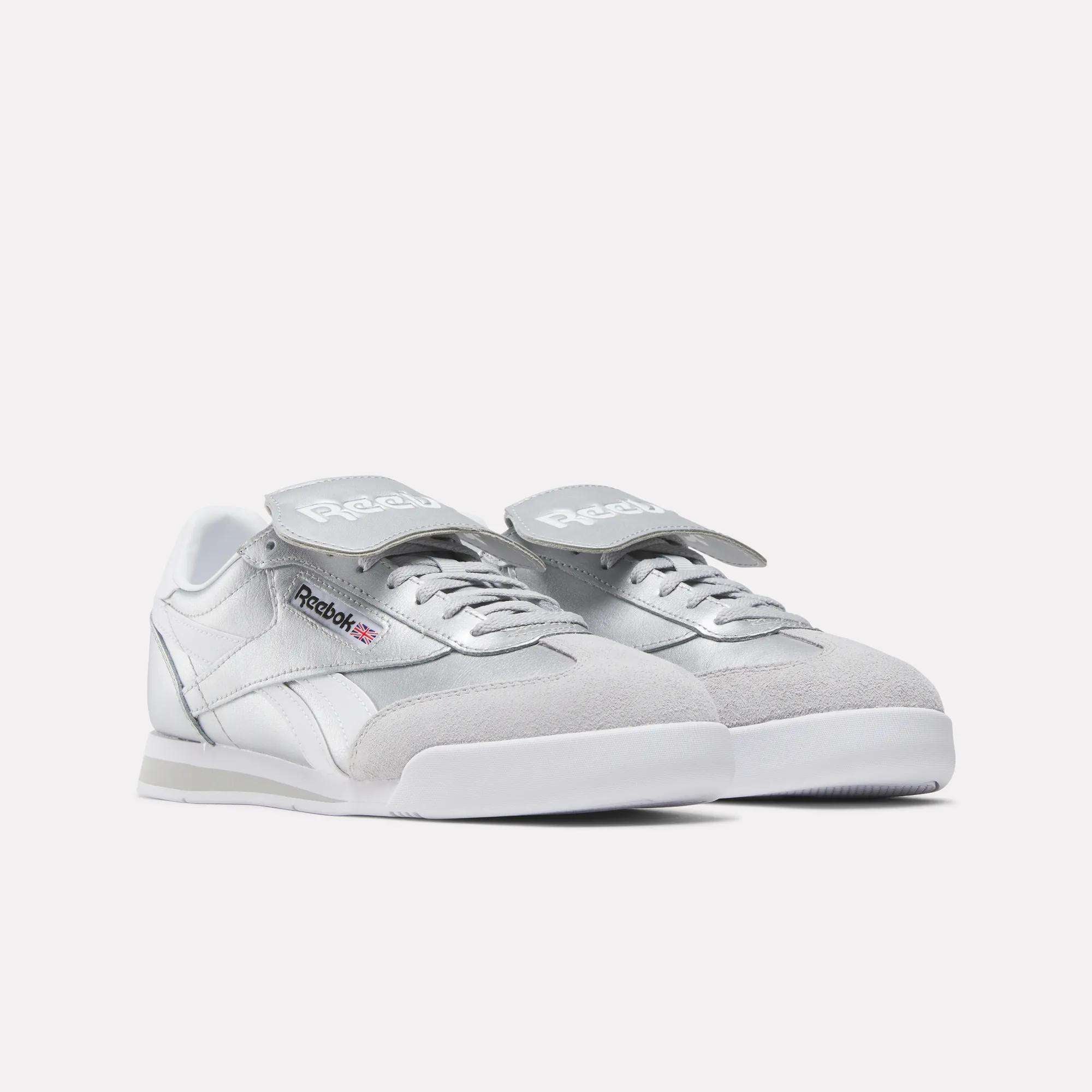A pair of Reebok Campio XT Kilty Shoes in light grey and white, with a classic tennis-inspired style, Velcro straps over the laces, and the Reebok logo on the side and tongue, shown on a plain white background.