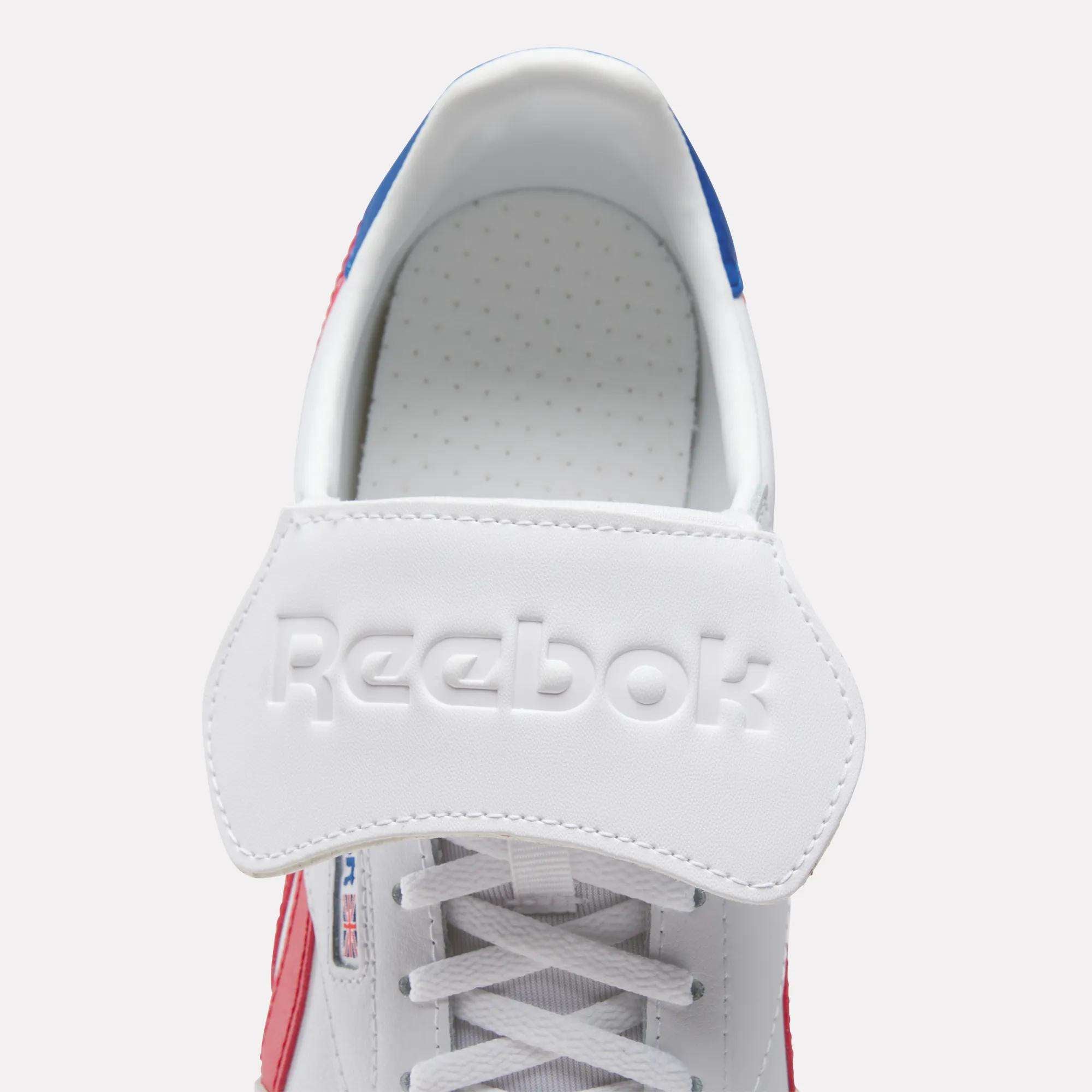 Top view of Reebok Campio XT Kilty Shoes in white, featuring a large branded strap with embossed logo on the tongue. This sleek, tennis-inspired sneaker includes red and blue accents and classic white laces.