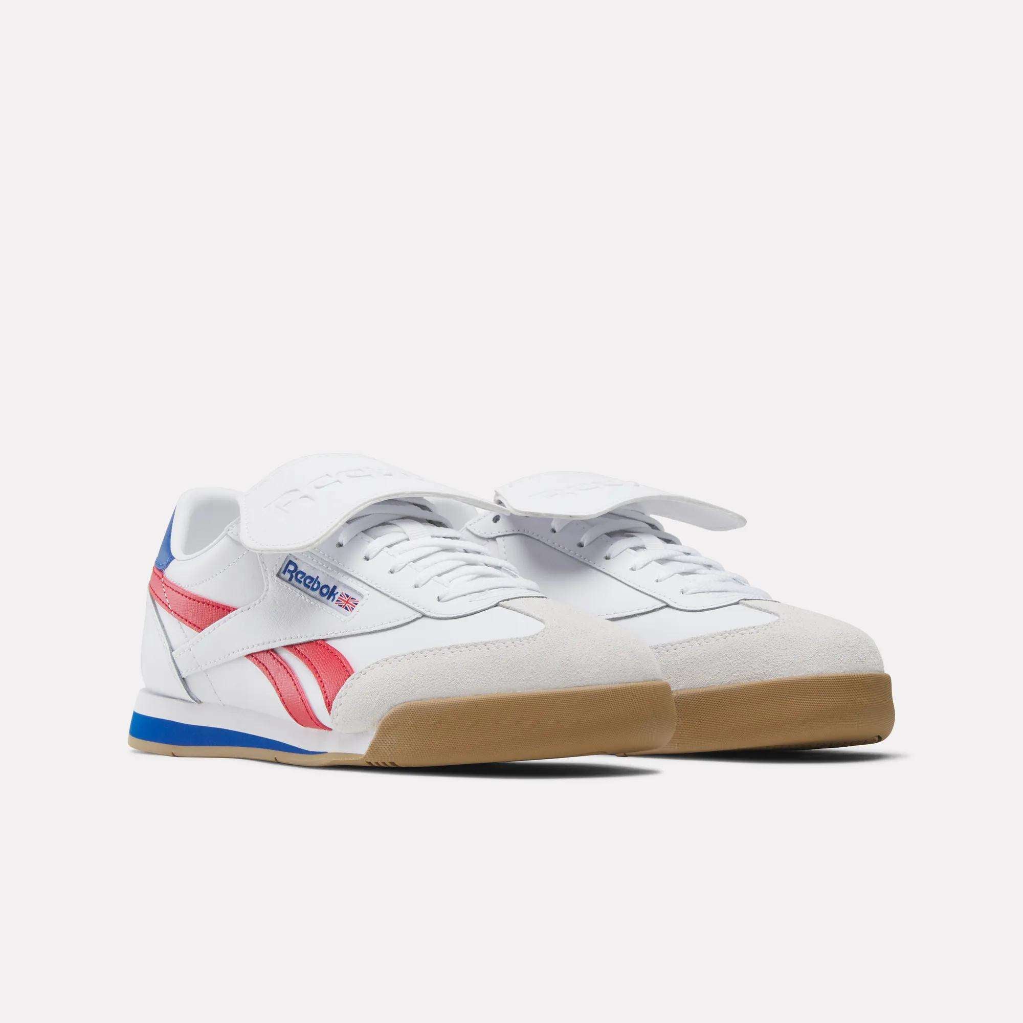 The Reebok Campio XT Kilty Shoes feature a white base with red and blue accents, beige suede toe detailing, gum soles, and a bold white strap over the laces, styled against a plain white background for tennis-inspired flair.