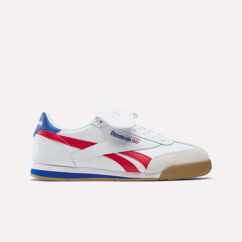 The Reebok Campio XT Kilty Shoes feature red stripes, blue accents, a tan suede toe cap, gum sole, and a Velcro strap on the tongue. Tennis-inspired with Reebok branding and UK flag on the side and heel.