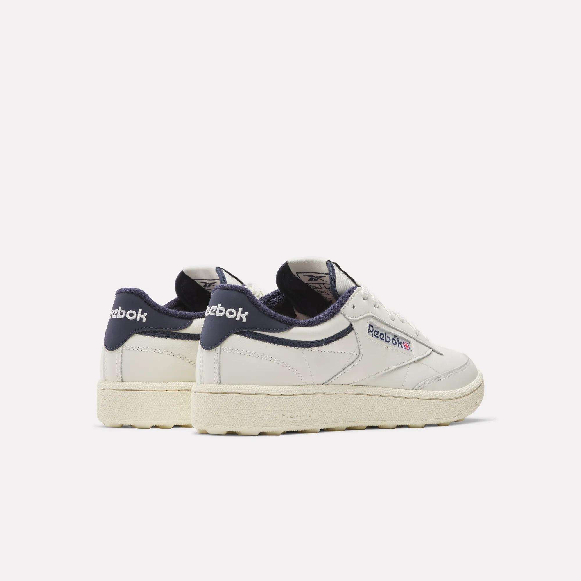 A pair of Reebok Men's Club C Golf Shoes in white with navy accents and off-white soles, featuring water-resistant leather uppers and a spikeless outsole, displayed on a plain light background, one showing the back and the other the side.