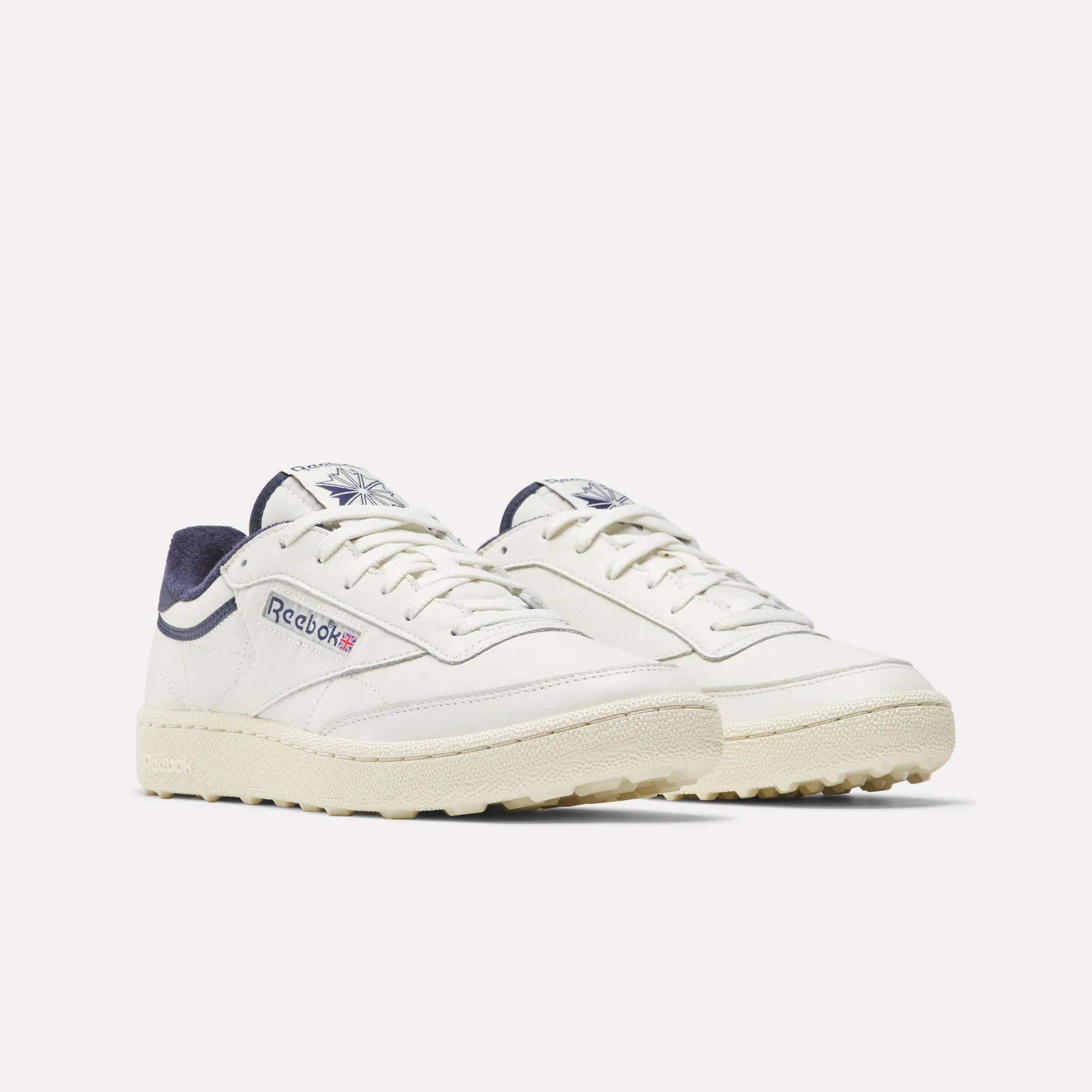 Men's Club C Golf Shoes by Reebok in off-white, featuring beige soles, navy accents on tongue and heel, side Reebok logo with Union Jack, and spikeless outsoles inspired by classic golf shoes.