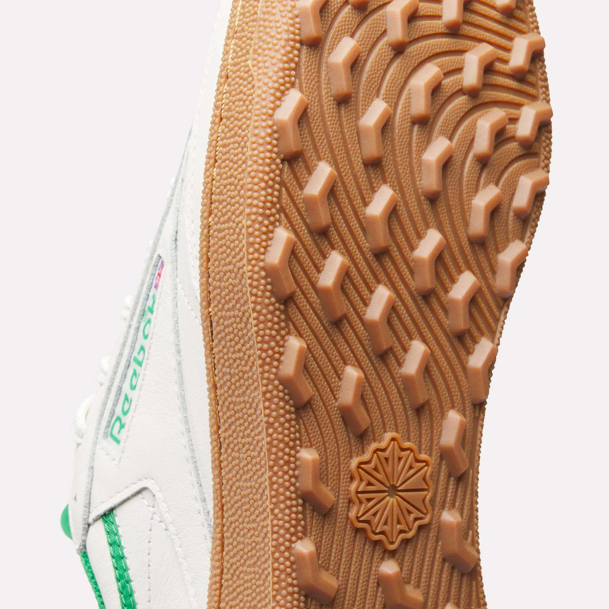Close-up of the sole on Reebok Men's Club C Golf Shoes. The gum-colored, spikeless outsole has a textured geometric tread and a central star-shaped emblem, with green accents highlighting the white sneaker.