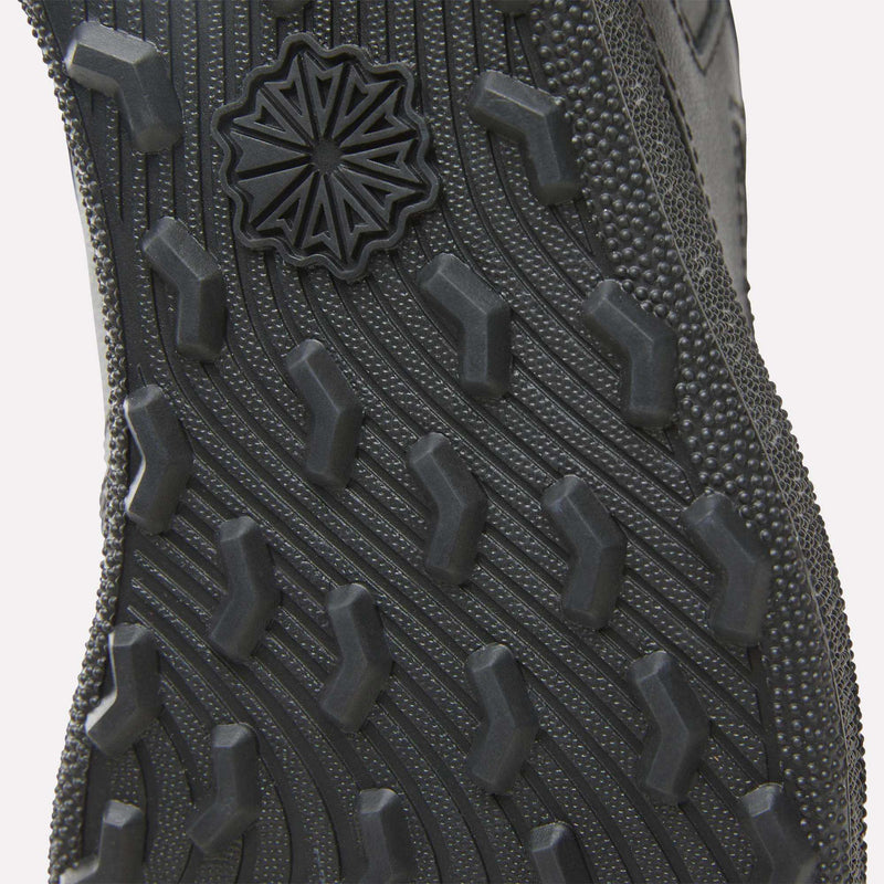 Close-up of the Reebok Men's Club C Golf Shoes in black, highlighting the spikeless sole with textured diagonal ridges, rectangular lugs, and a starburst design near the toe.