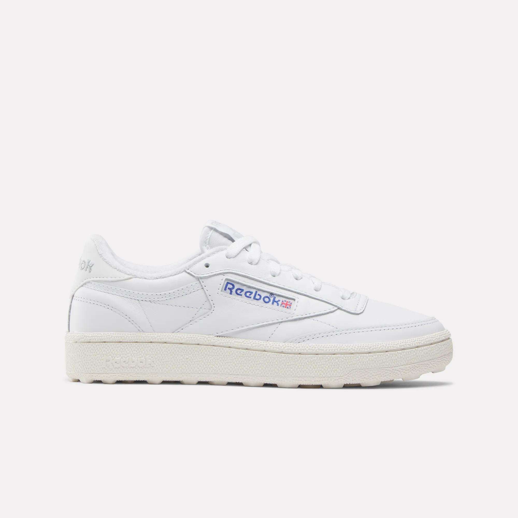 The Reebok Women’s Club C Golf Shoes feature a white upper, cream sole, blue and red side logo, spikeless outsole, and white laces, shown against a plain white background.