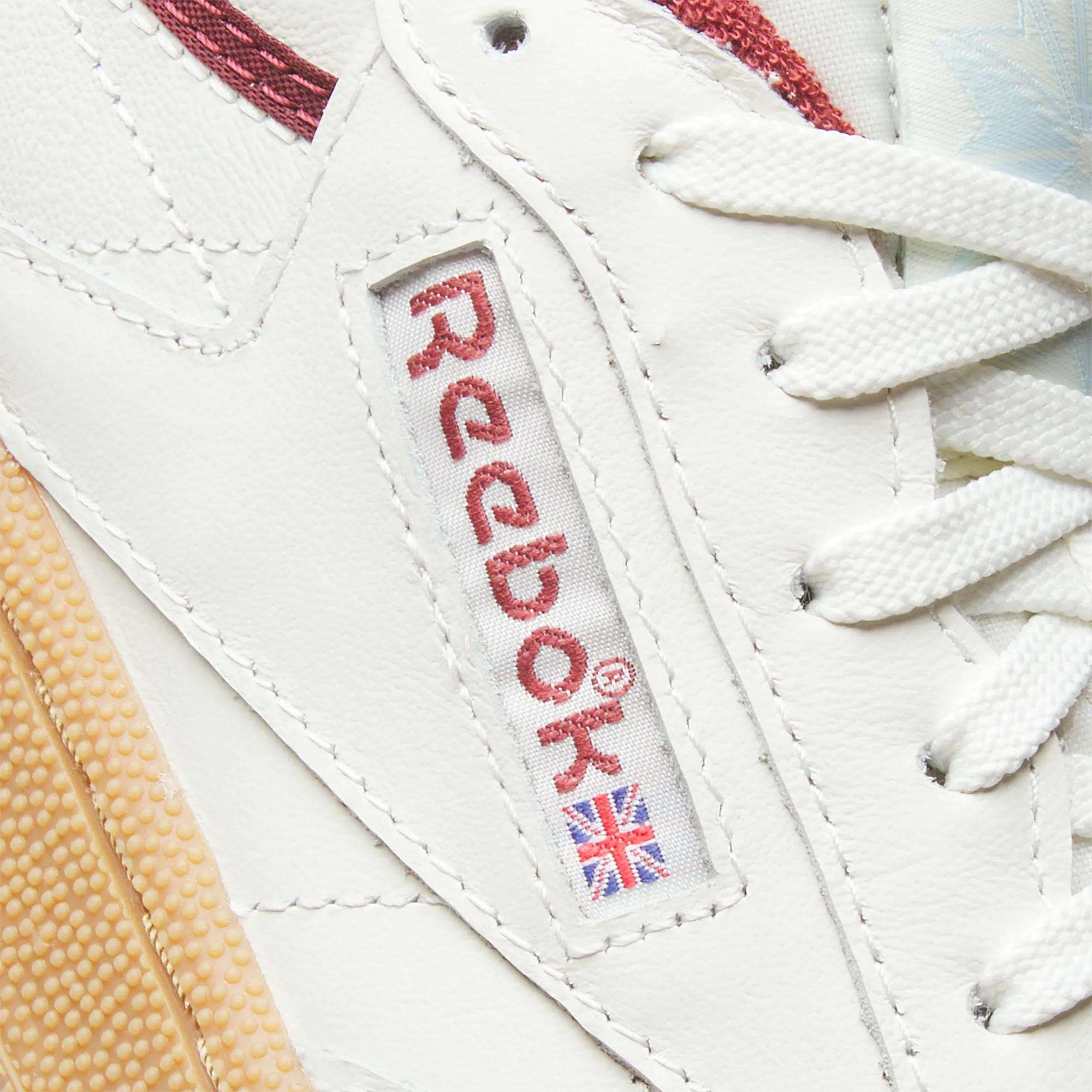 Close-up of Reebok Women's Club C Golf Shoes, showing white water-resistant leather, a stitched red logo with Union Jack flag, white laces, and a beige textured sole for enhanced durability.