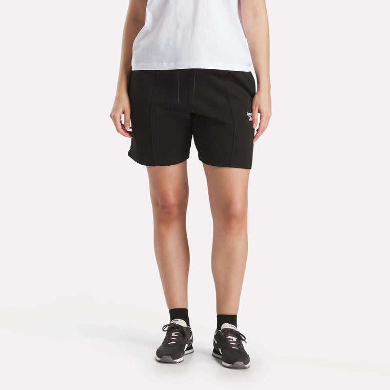 A person faces forward against a plain white background, wearing a white t-shirt, Reebok Women's Pintuck Long Shorts, black socks, and black sneakers with their hands relaxed at their sides.