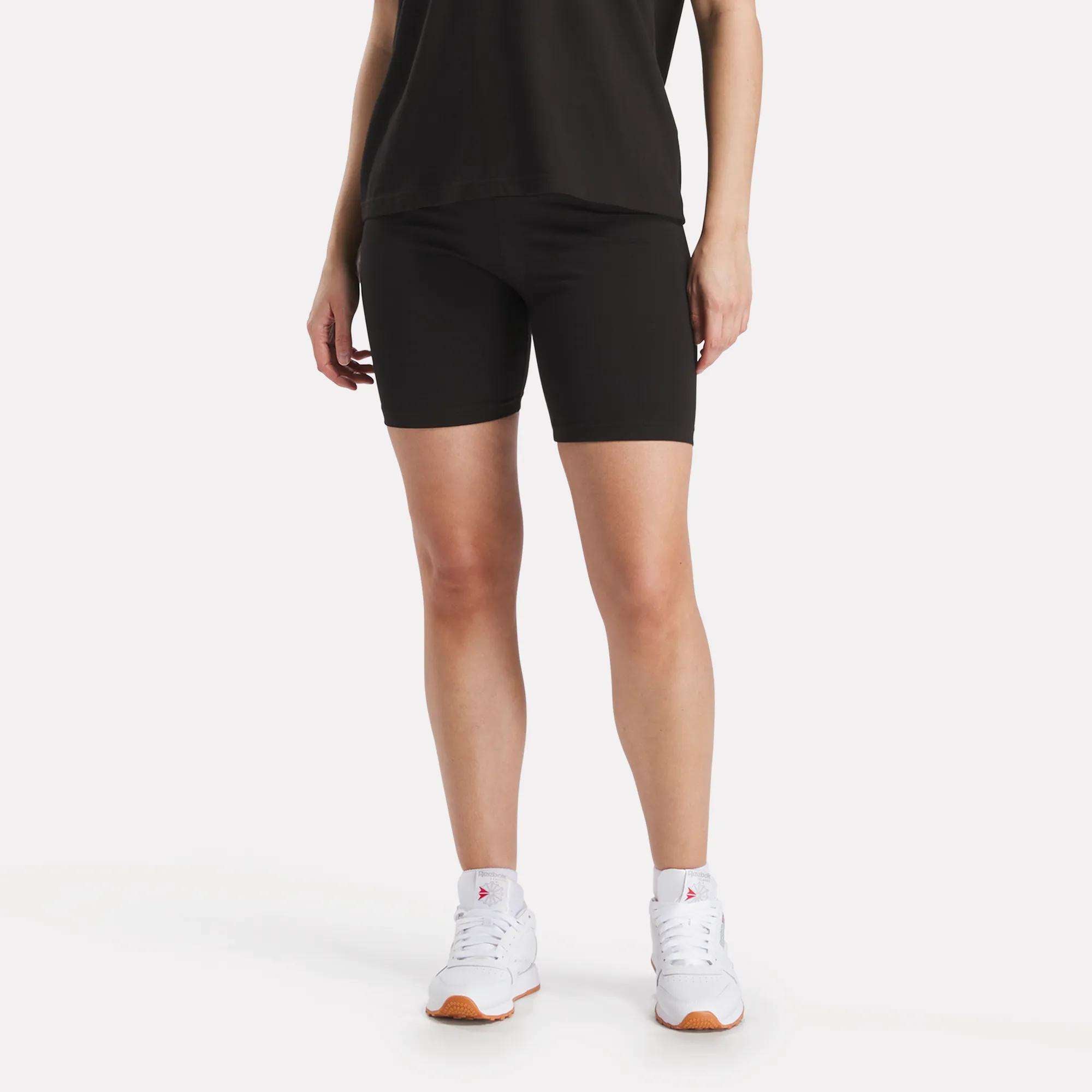 A person wears Reebok Women's Small Logo Cycle Shorts, a black T-shirt, and white sneakers against a plain white background. Only the lower half of the body is visible.