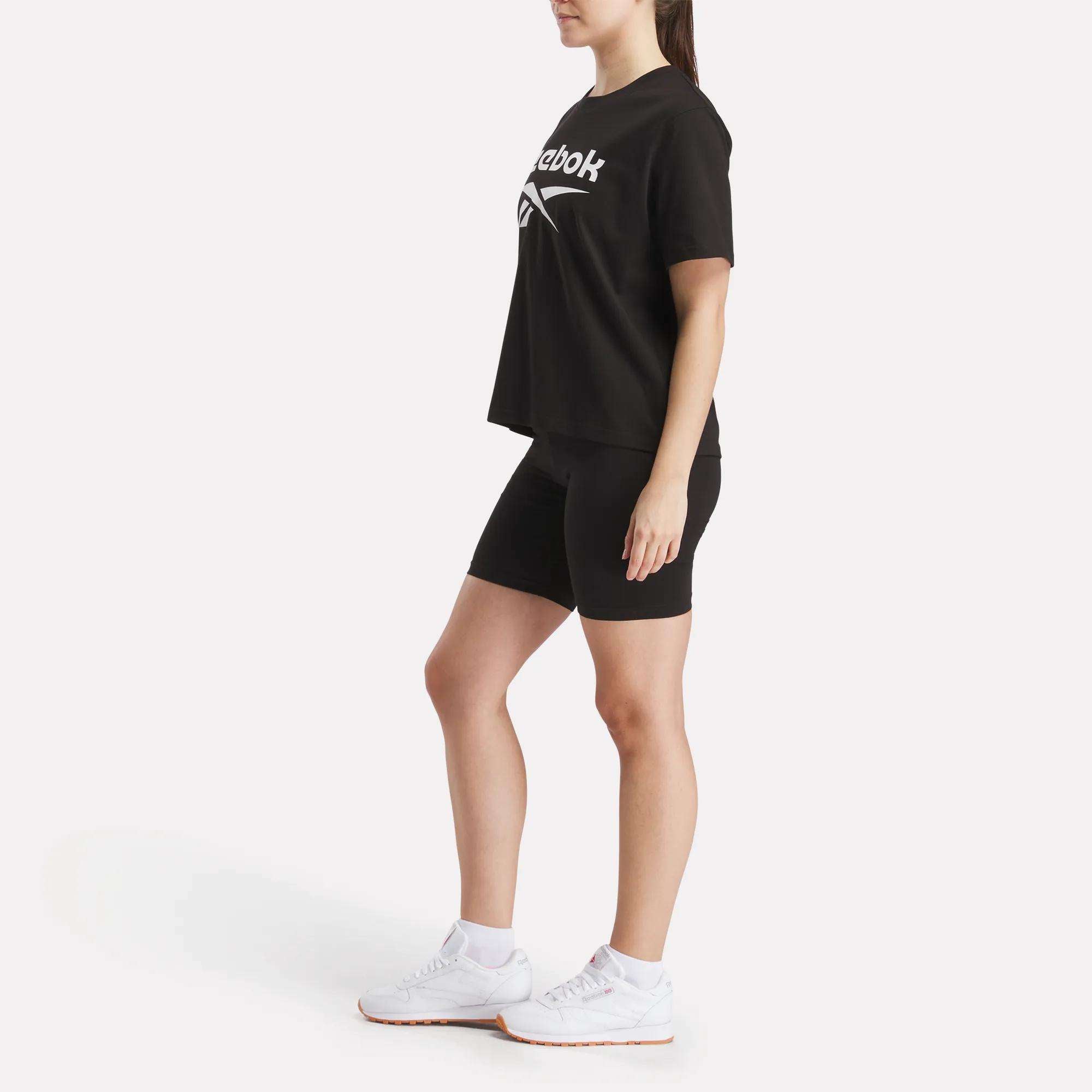 A person stands on a white background wearing a black Reebok t-shirt, Women's Reebok Small Logo Cycle Shorts with a stretchy fit, and white sneakers, facing slightly to the side with arms relaxed at their sides.