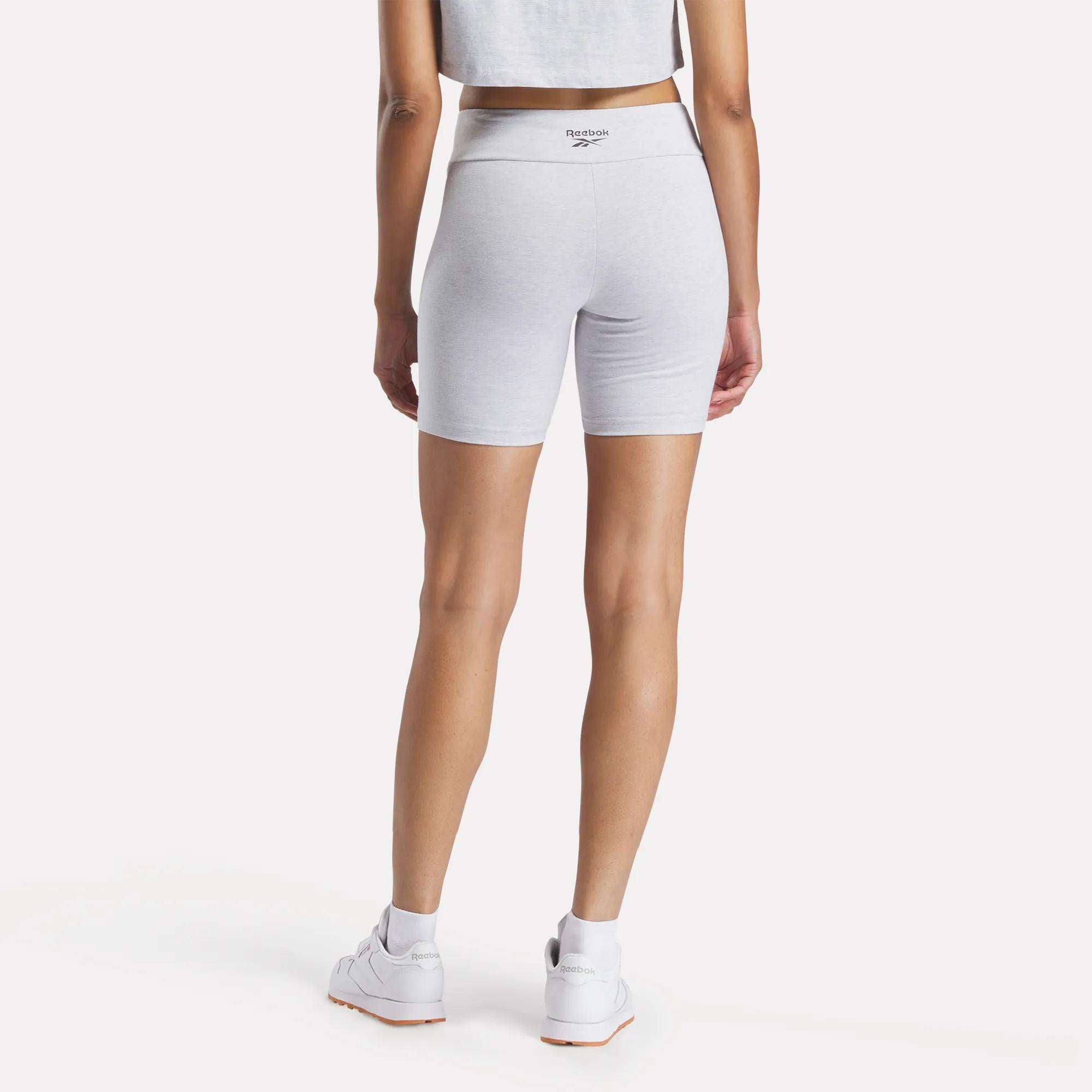 A woman in a gray crop top, Reebok Women's Small Logo Cycle Shorts in light gray, and white Reebok sneakers is photographed from behind against a plain white background.