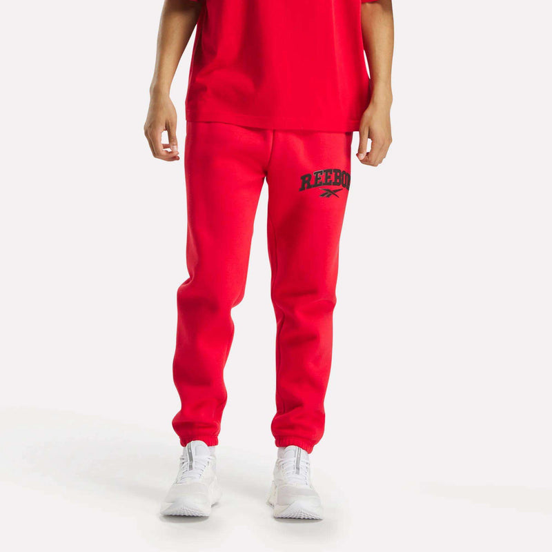 A man models Reebok Men's Joggers in red, featuring a black logo, against a white background. Only the lower half is shown, paired with white sneakers.