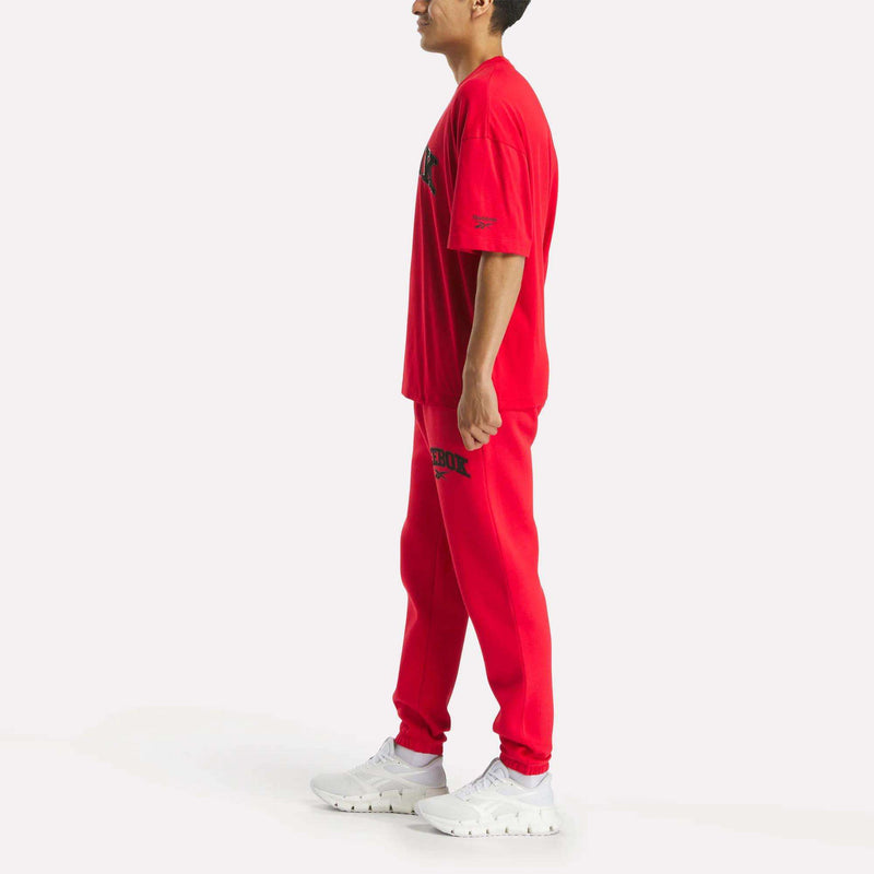 A person stands sideways against a plain white background wearing Reebok Men's Joggers and a matching bright red T-shirt, both featuring black text, paired with white sneakers.