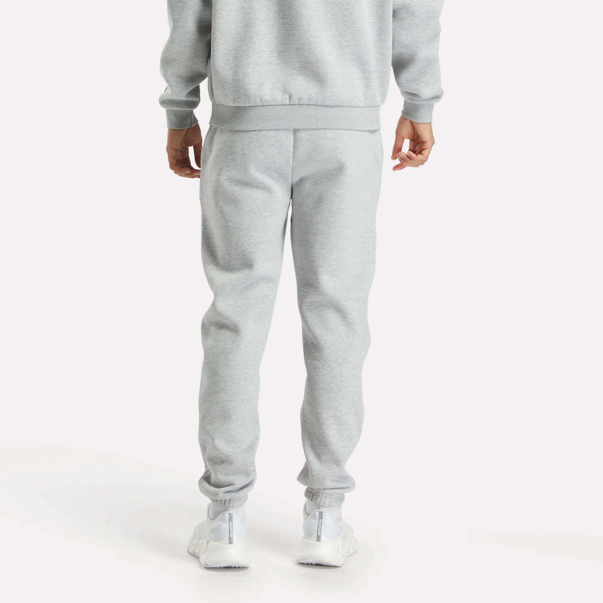 A person faces away, wearing Reebok Men's Joggers in light gray with a matching sweatshirt and white sneakers, set against a plain white background.