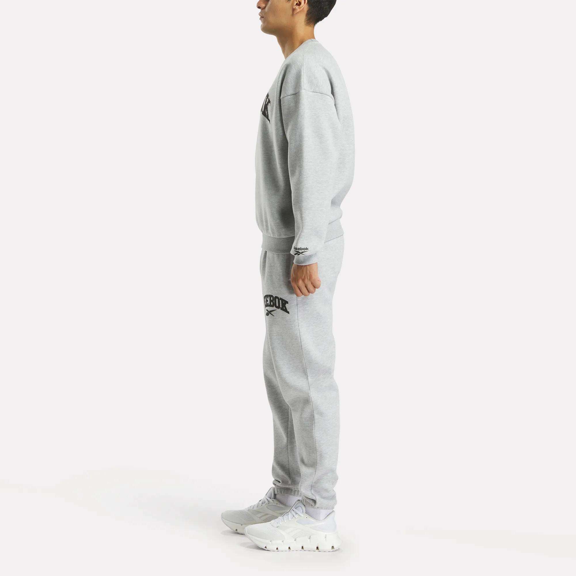 A person stands in profile wearing Reebok Men's Joggers with a matching light gray sweatshirt and white sneakers, set against a plain white background.