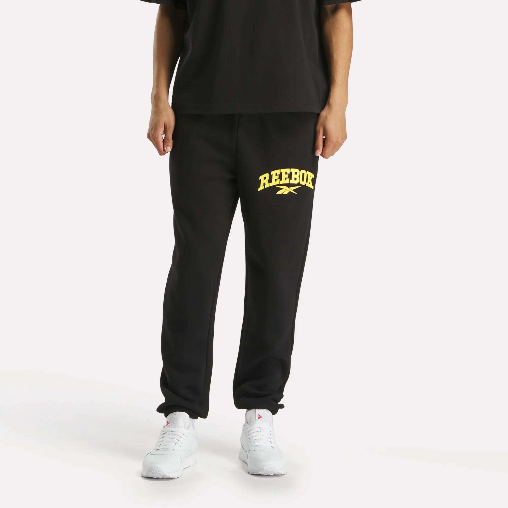 A person wearing Reebok Men's Joggers in black with a yellow logo on the left thigh, paired with a black T-shirt and white sneakers, stands against a plain white background.