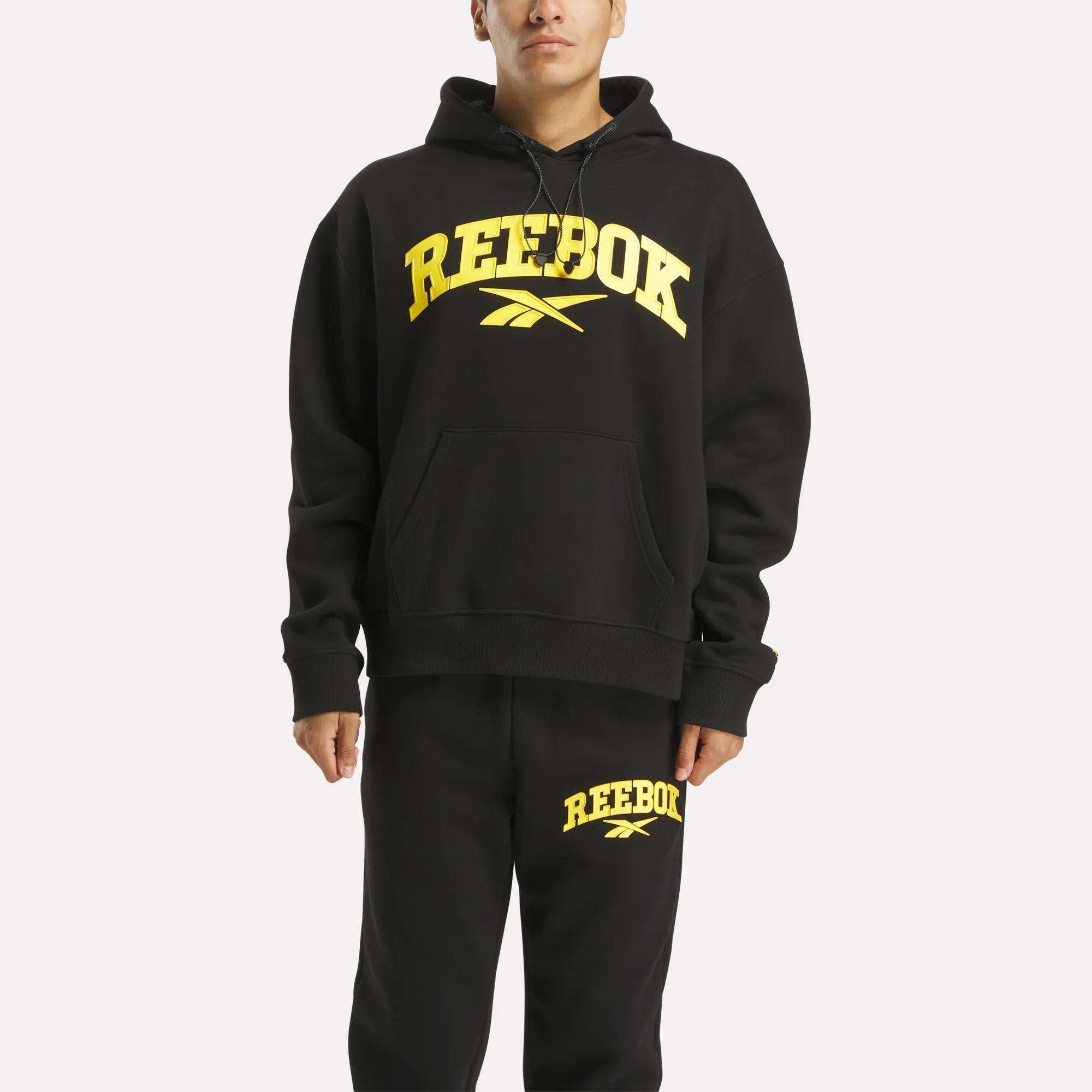 A person stands against a white background wearing a black Reebok Men's Hoodie and matching black sweatpants, both with bold yellow Reebok logos and text. The hoodie offers comfort and effortless style.