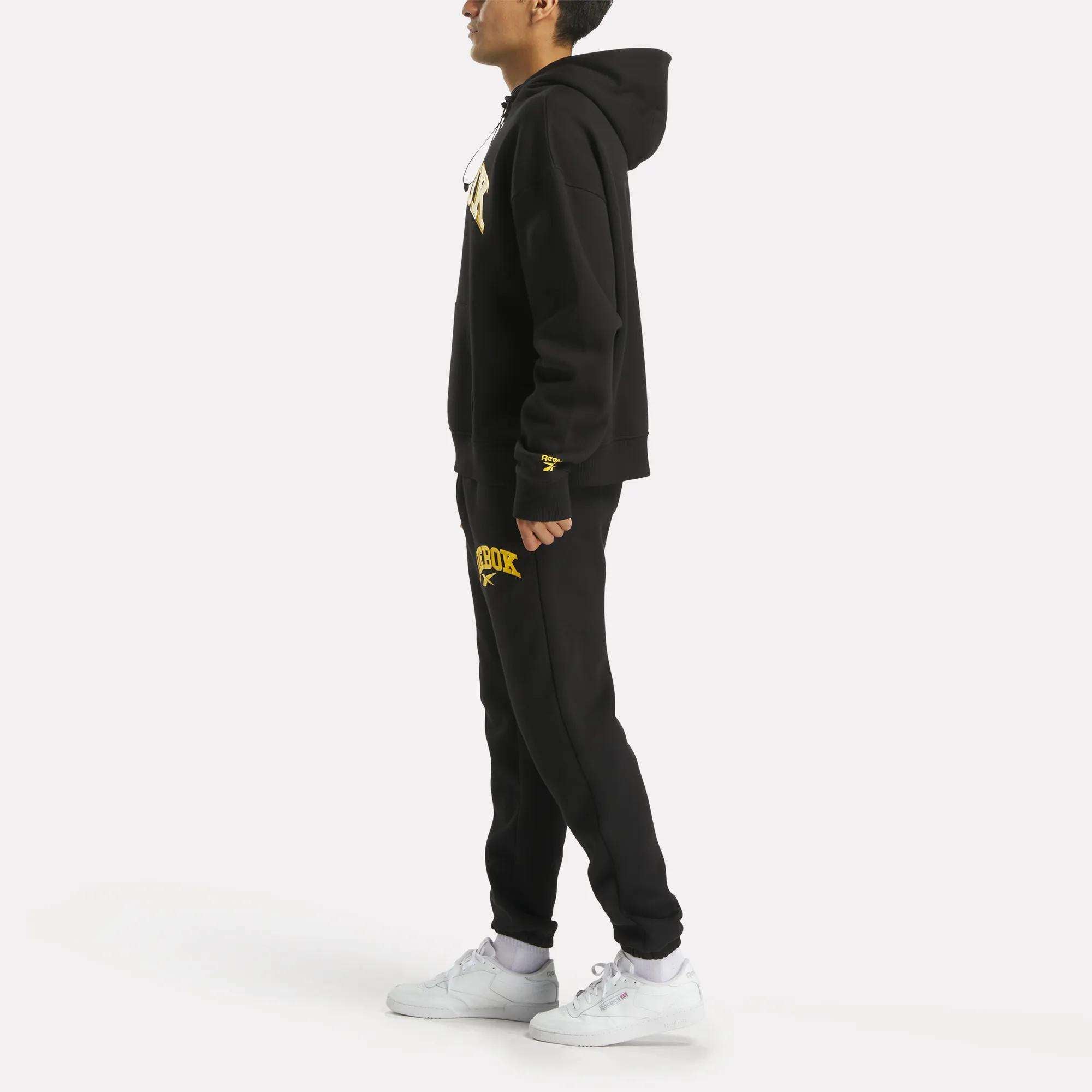 A man in profile wears a Reebok Men’s Hoodie in black and matching black sweatpants with yellow lettering, paired with white sneakers, set against a plain white background.