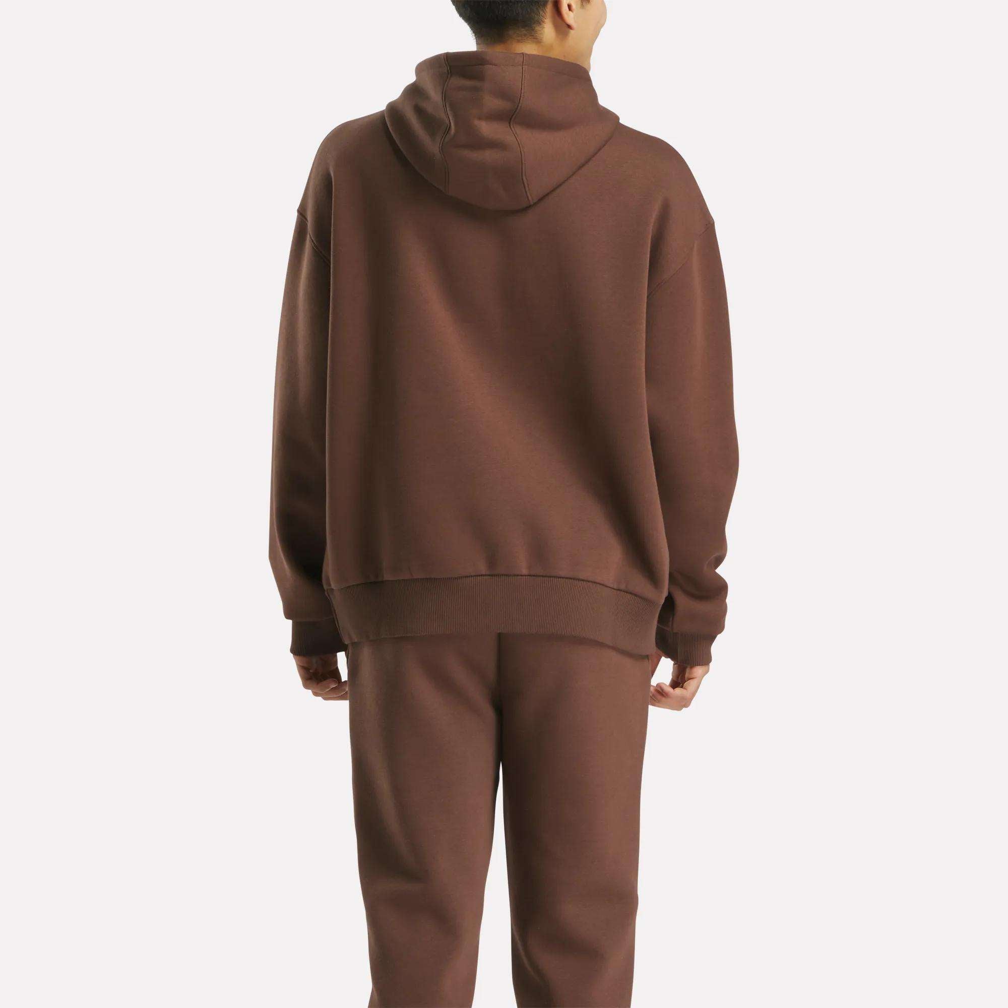 A person stands with their back to the camera, wearing a Reebok Men's Varsity Hoodie in brown, paired with matching sweatpants against a plain light background.