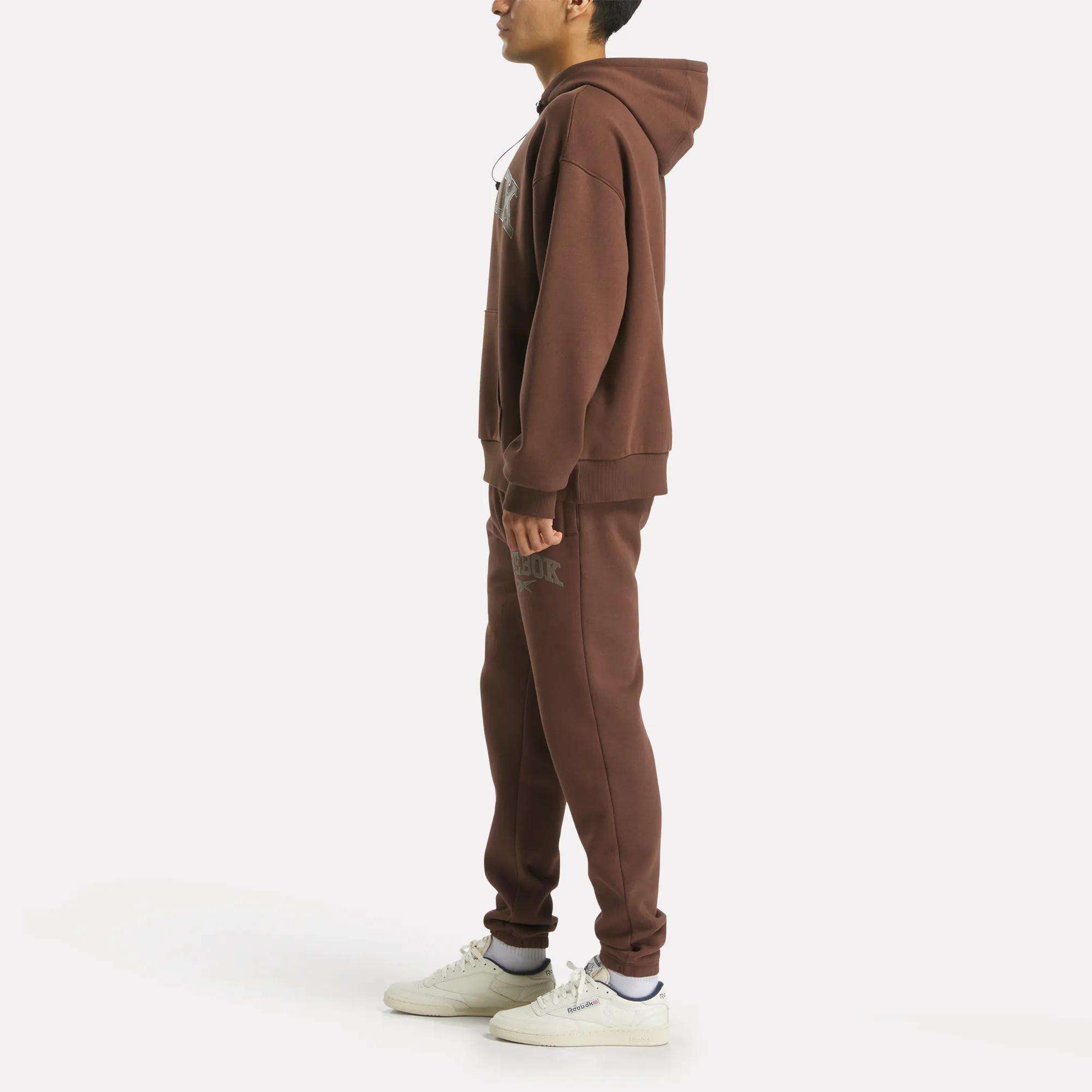 A man stands in profile wearing the Reebok Men's Varsity Hoodie in brown with matching sweatpants and white sneakers, capturing a relaxed collegiate vibe against a plain white background.