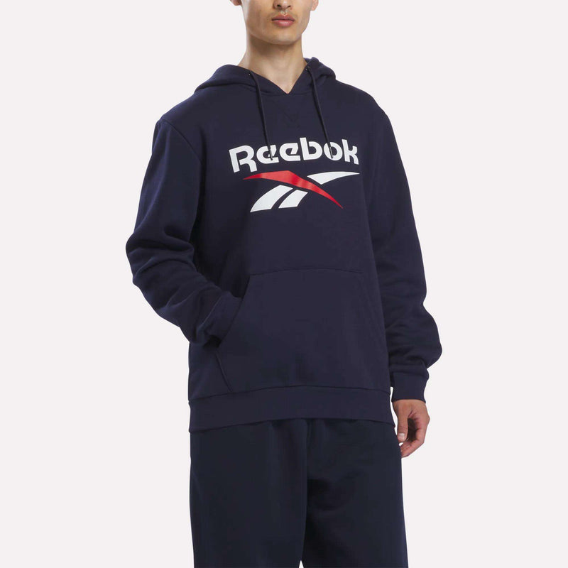A man stands against a plain background with one hand in the pocket, wearing the Reebok Men’s Logo Hoodie—a navy blue cotton-blend fleece hoodie featuring a bold white and red Reebok logo on the front.