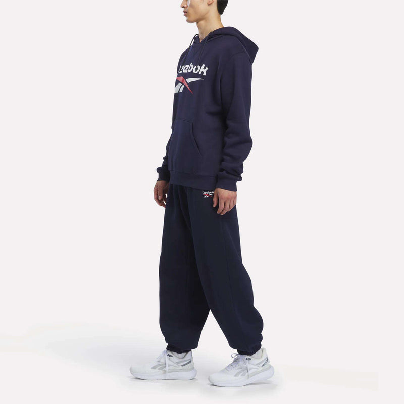 A man is wearing a Reebok Men’s Logo Hoodie in navy, crafted from cotton-blend fleece, paired with matching sweatpants and white sneakers, standing against a plain white background.