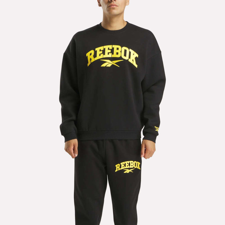 A person wears a black Reebok Men's Cottle Sweatshirt with bold yellow logos, paired with matching sweatpants, creating a sporty, casual look against the plain white background.