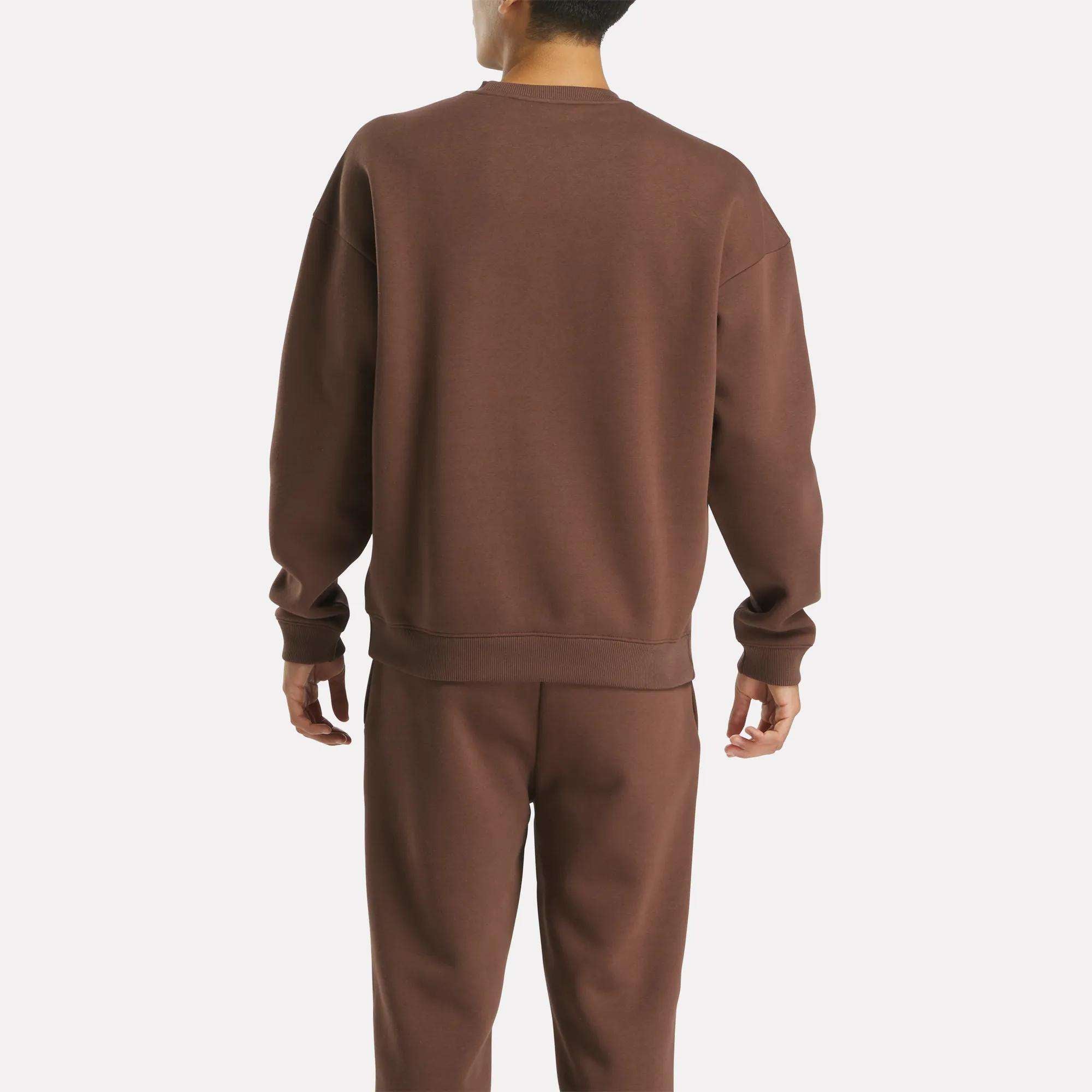 A person faces away from the camera, wearing the Reebok Men's Varsity Sweatshirt in brown with matching sweatpants, set against a plain white background.