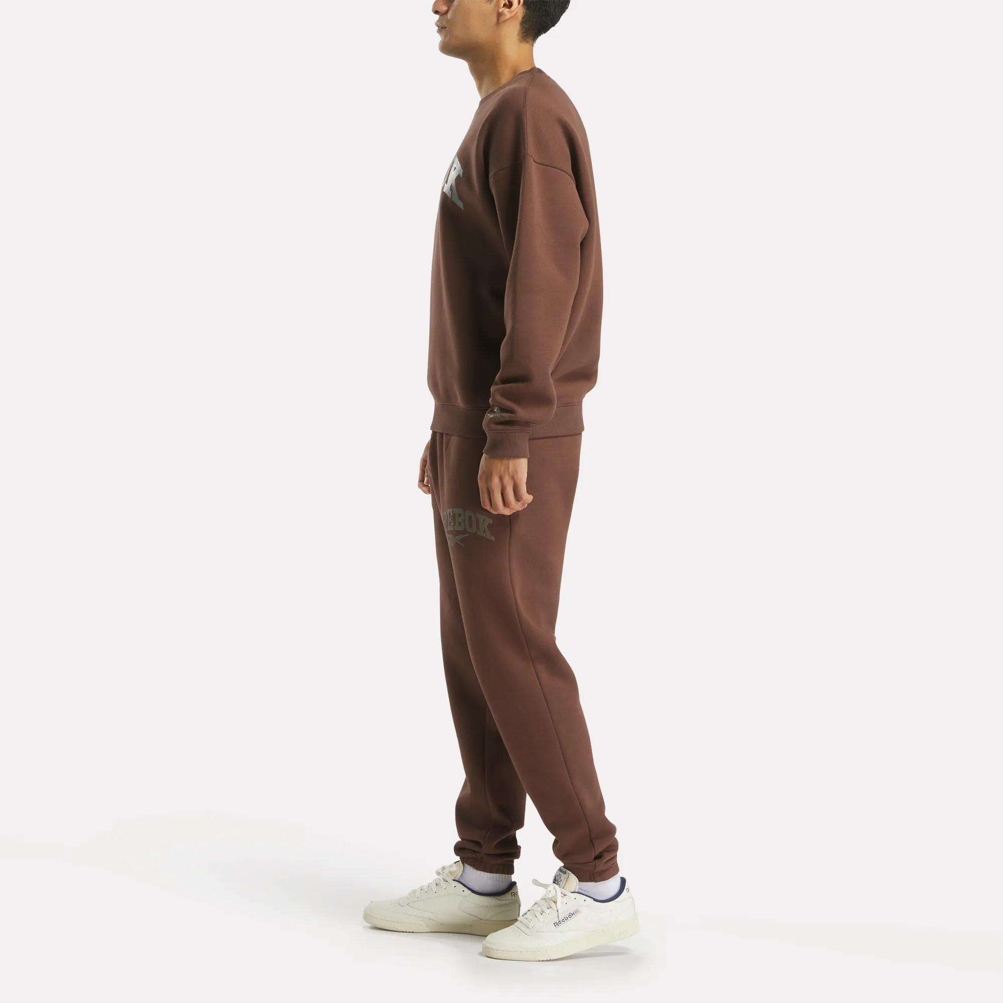 A man stands sideways in a matching brown Reebok Men's Varsity Sweatshirt and sweatpants with white sneakers, set against a plain white backdrop.