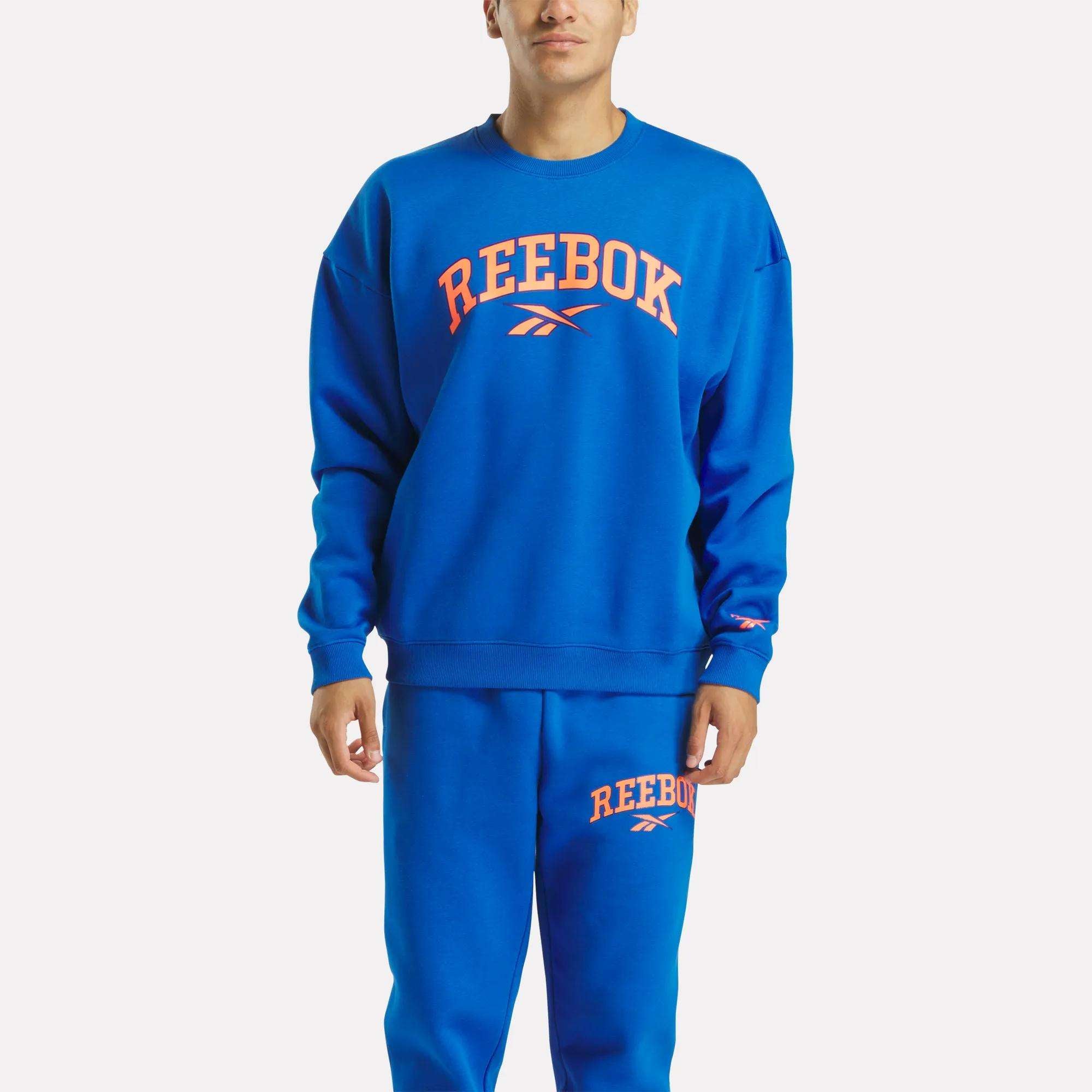 A person wears a blue Reebok Men's Varsity Sweatshirt featuring bold orange REEBOK branding, standing against a plain light background.