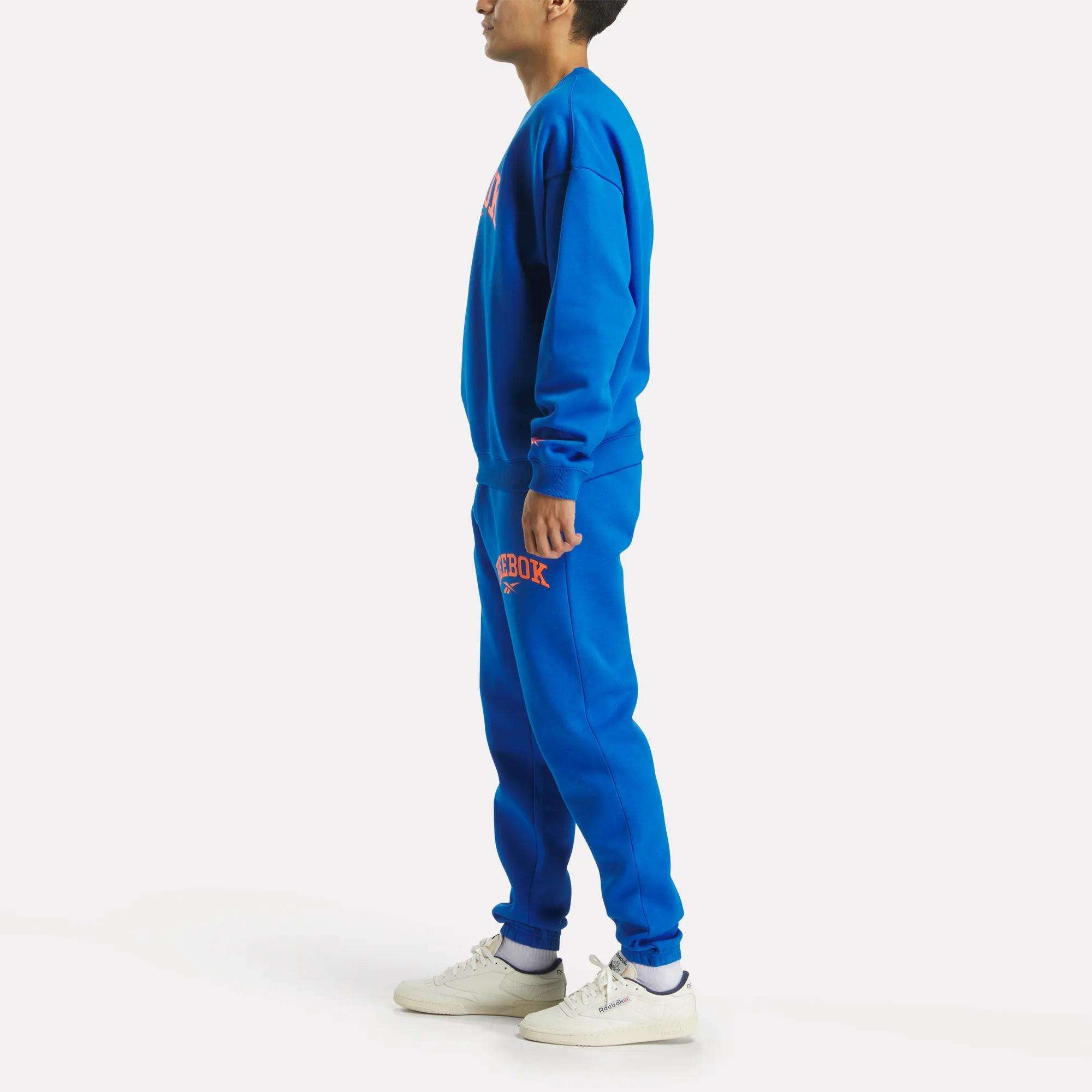 A man in a bright blue Reebok Men's Varsity Sweatshirt and sweatpants set with bold logos stands sideways in white sneakers against a plain white background.