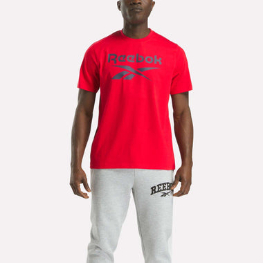 A man faces forward against a plain white background, dressed in a bright red Reebok Men's Big Logo Crewneck Short Sleeve Tee and light gray Reebok sweatpants, both featuring prominent brand logos.