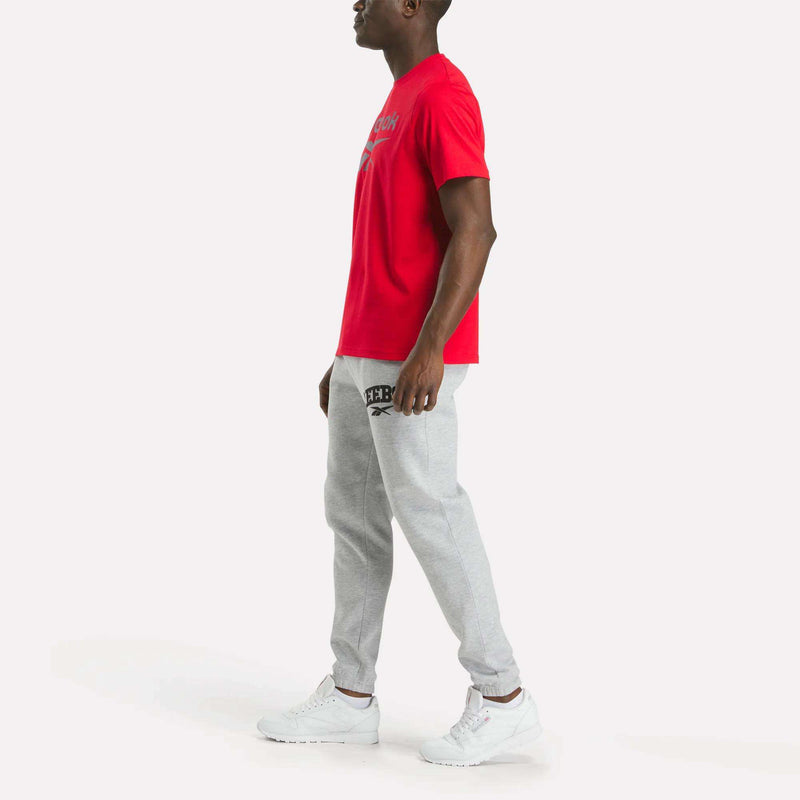 A man stands sideways wearing a Reebok Men's Big Logo Crewneck Short Sleeve Tee in red, light gray sweatpants, and white sneakers against a plain white background.