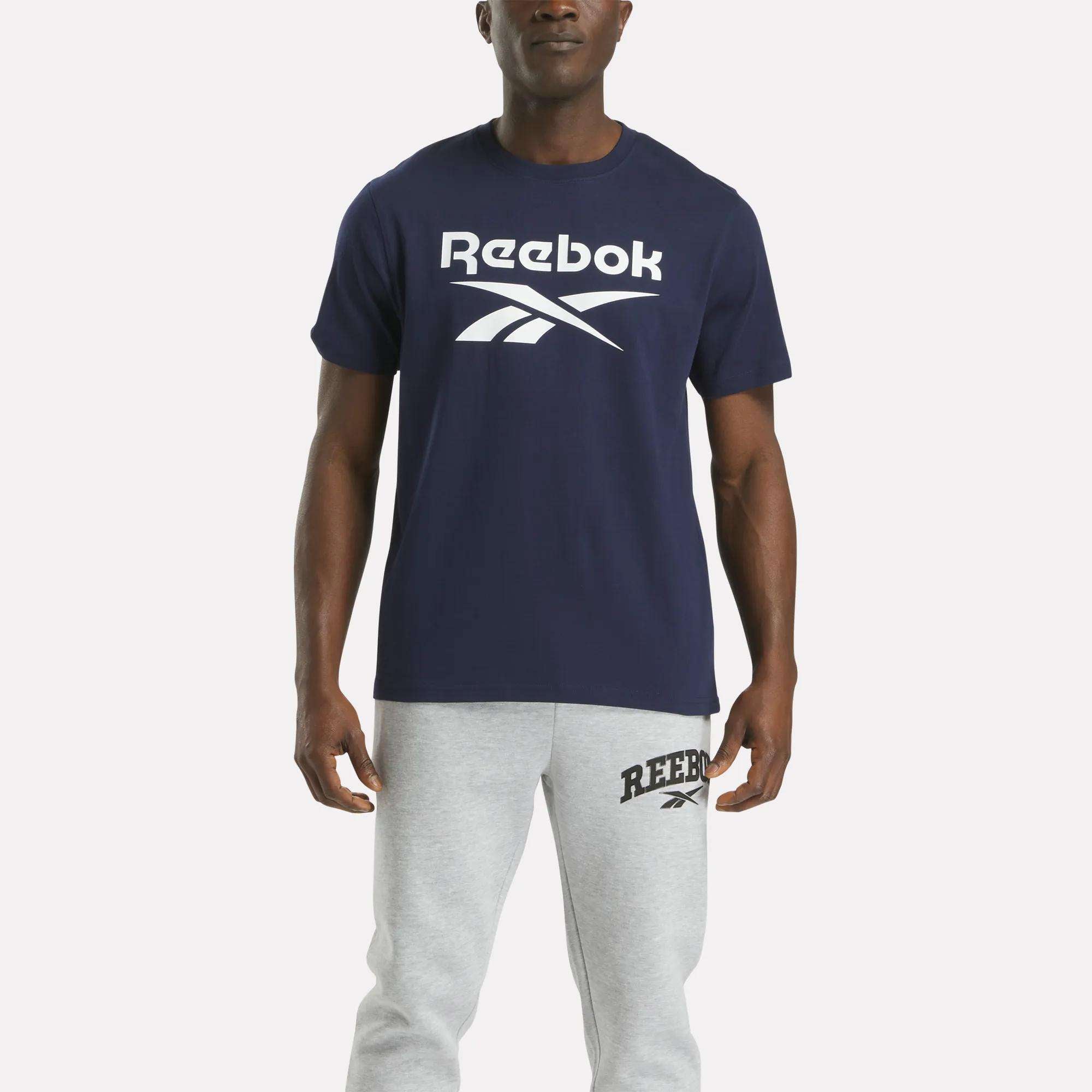 A person faces forward wearing the Reebok Men's Big Logo Crewneck Short Sleeve Tee in navy with a large white logo, paired with light gray sweatpants featuring a small logo near the left pocket.