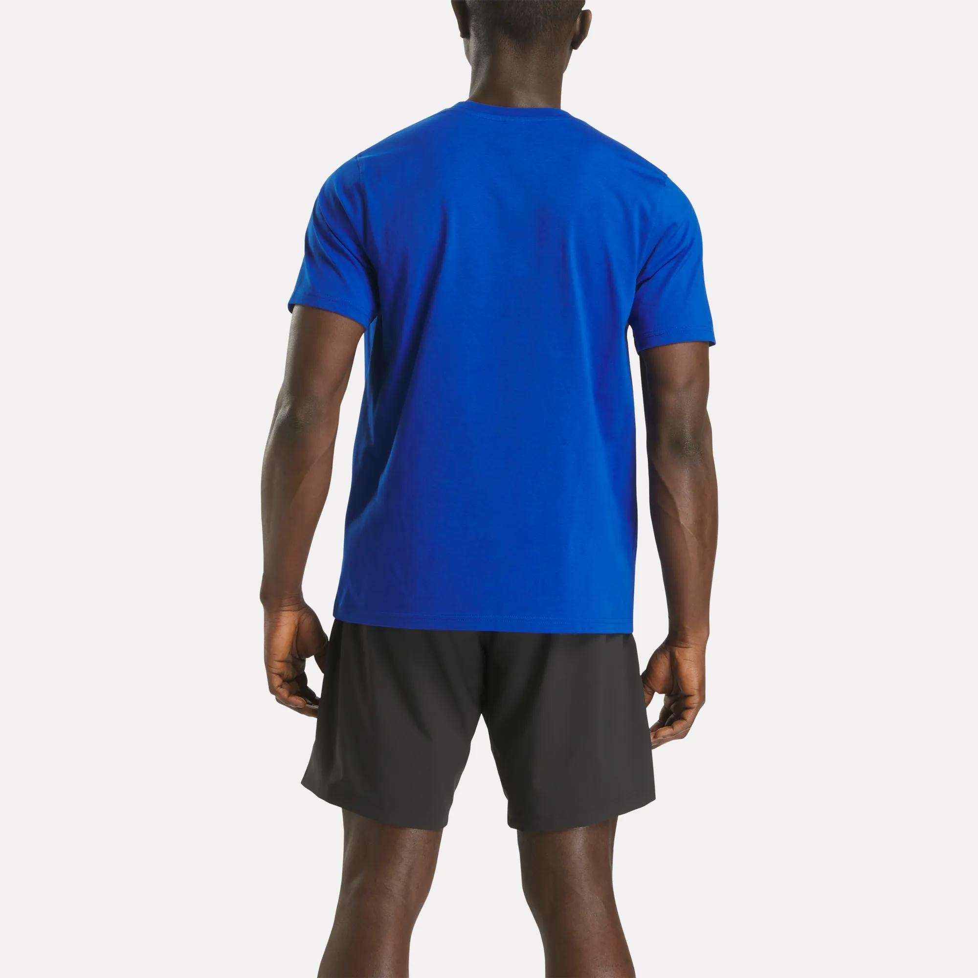 A man faces away from the camera, wearing a Reebok Men's Small Logo Crewneck Short Sleeve T-Shirt in bright blue and black athletic shorts against a plain white background.