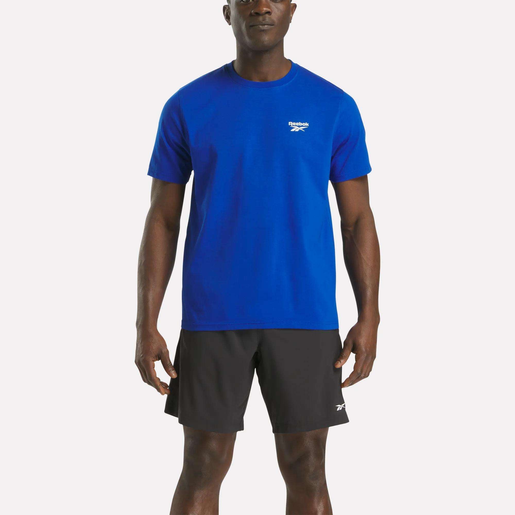 A man faces forward wearing a Reebok Men's Small Logo Crewneck Short Sleeve T-Shirt in blue and black Reebok shorts against a plain white background.