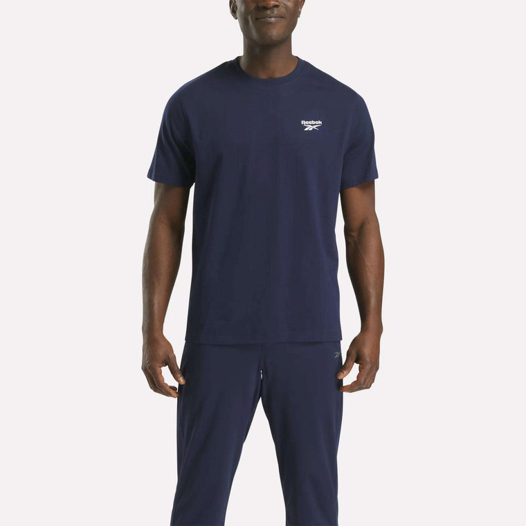 A man in a navy blue Reebok Men's Small Logo Crewneck Short Sleeve T-Shirt stands facing the camera against a plain white background. This classic fit tee features a small Reebok logo on the left chest.