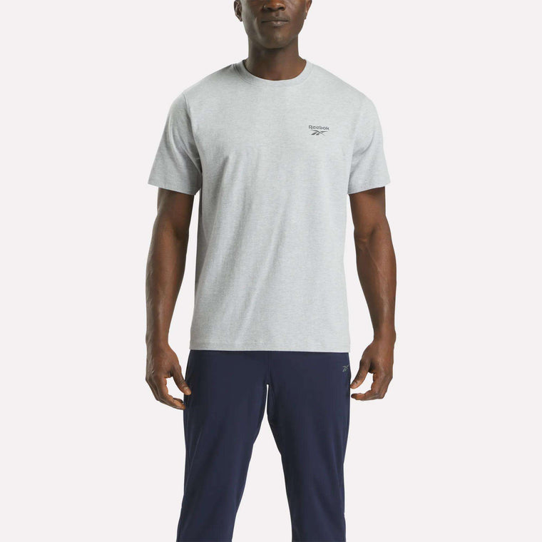 A man faces forward, wearing a Reebok Men's Small Logo Crewneck Short Sleeve T-Shirt in light grey cotton jersey and dark navy pants, set against a plain white background.