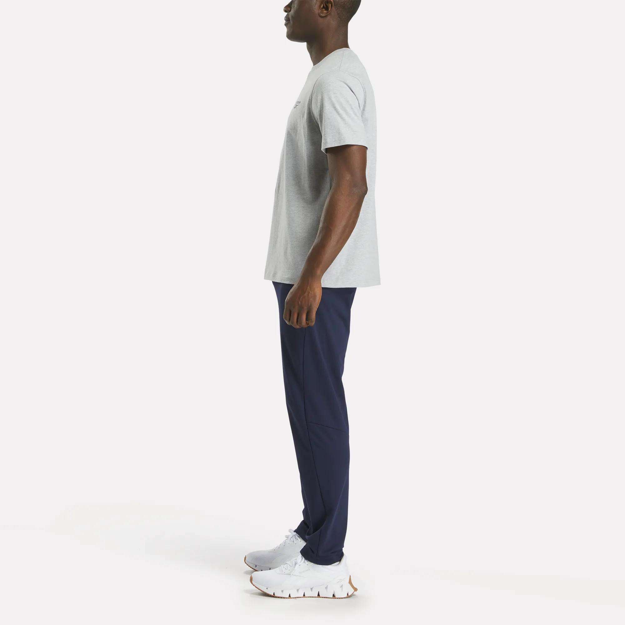 A man stands in profile facing left, wearing a Reebok Men's Small Logo Crewneck Short Sleeve T-Shirt in light gray cotton jersey, paired with navy blue athletic pants and white sneakers, against a plain light background.