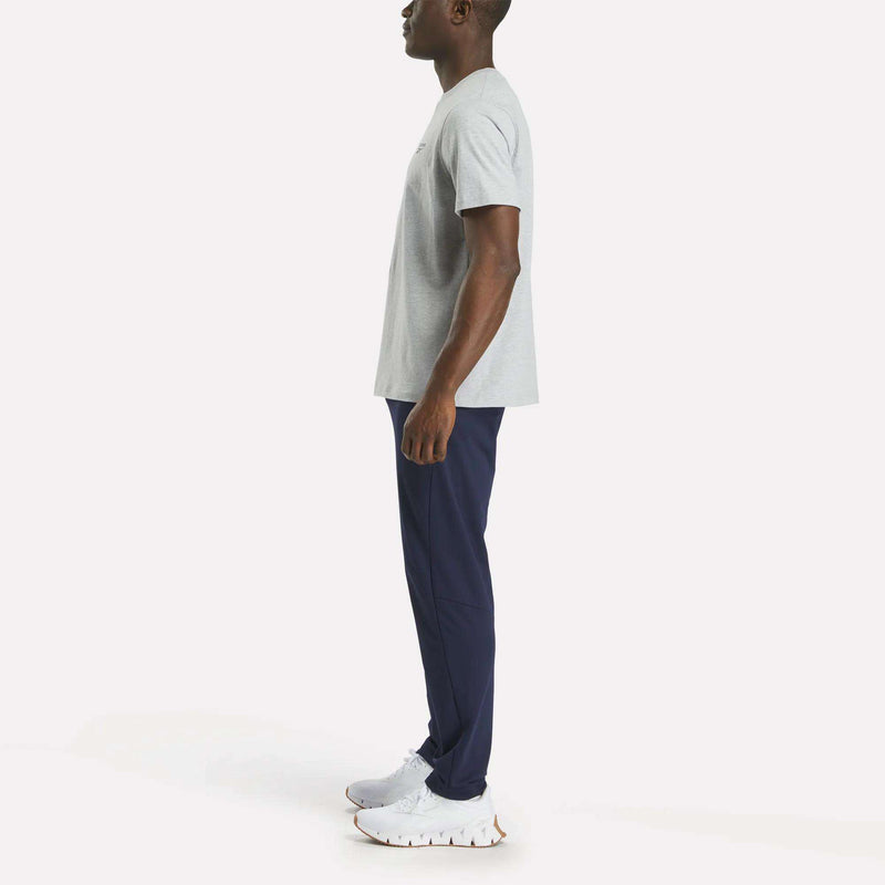 A man stands in profile facing left, wearing a Reebok Men's Small Logo Crewneck Short Sleeve T-Shirt in light gray cotton jersey, paired with navy blue athletic pants and white sneakers, against a plain light background.